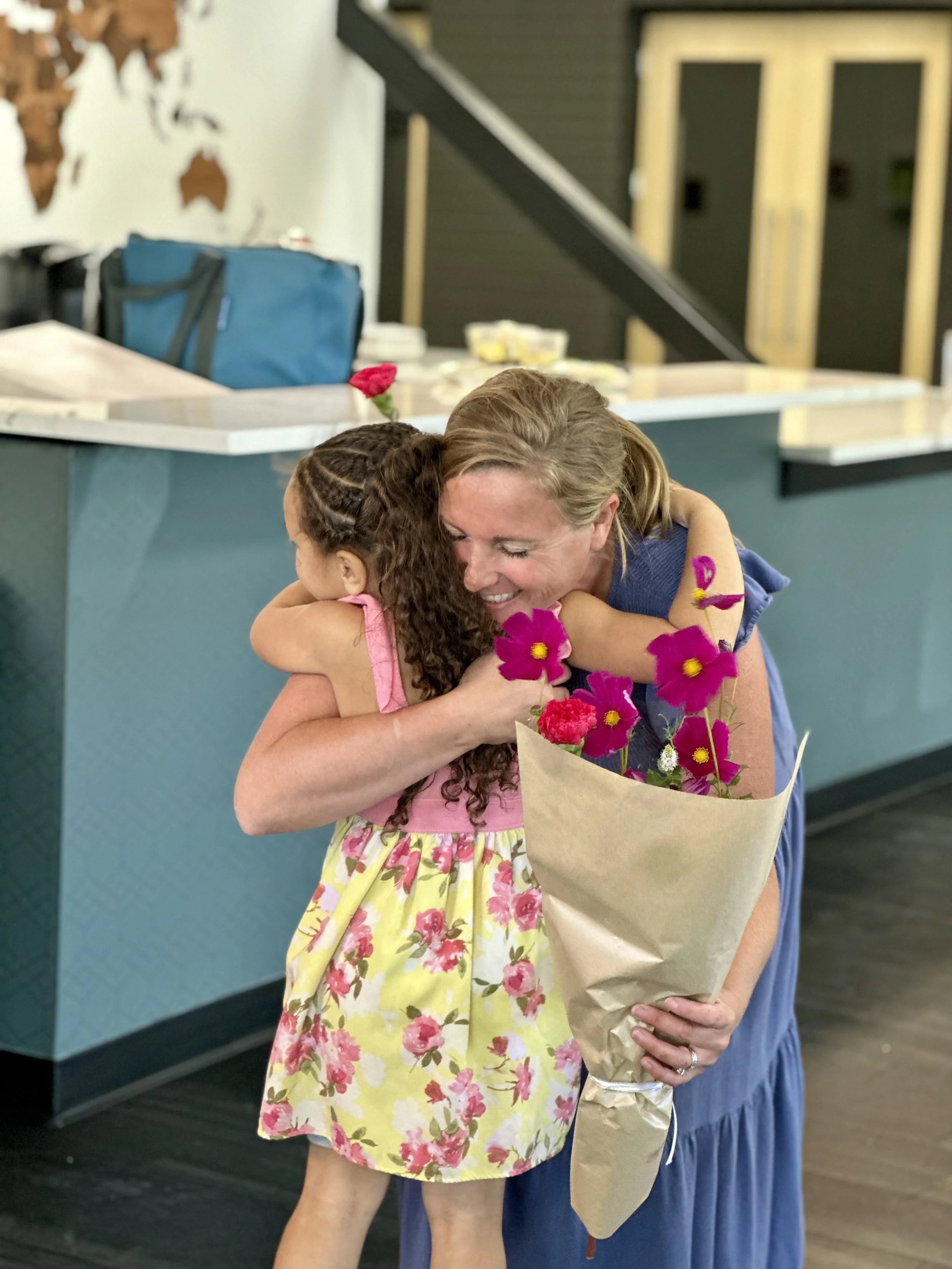 A woman and a girl hugging warmly; the woman is holding a bouquet of pink flowers.