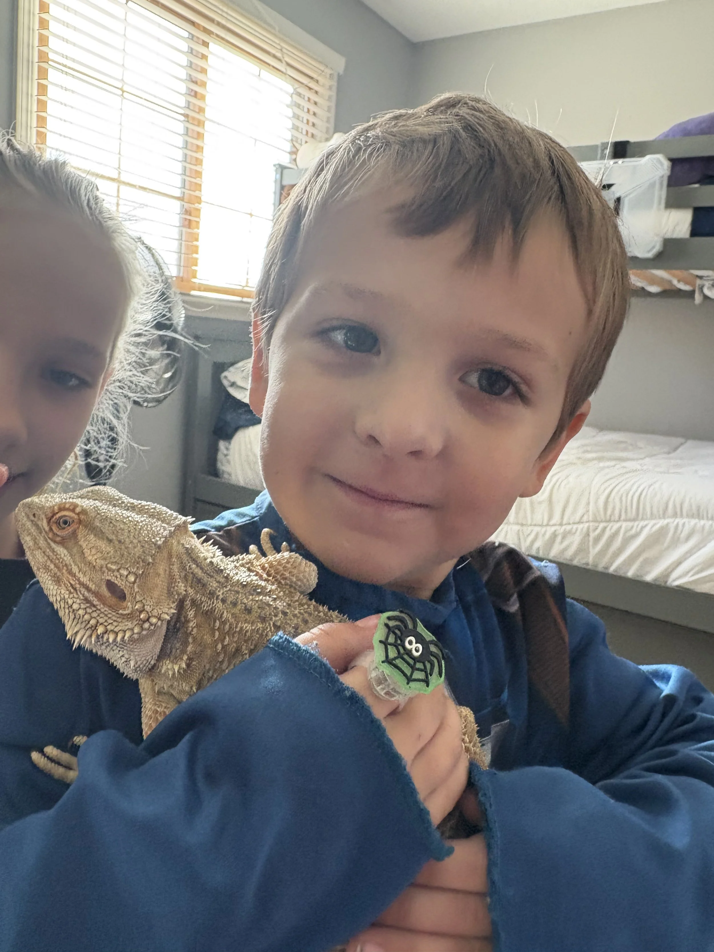 A young boy holding a bearded dragon lizard, with another child partially visible on the left, inside a room with a bed and a window with blinds.