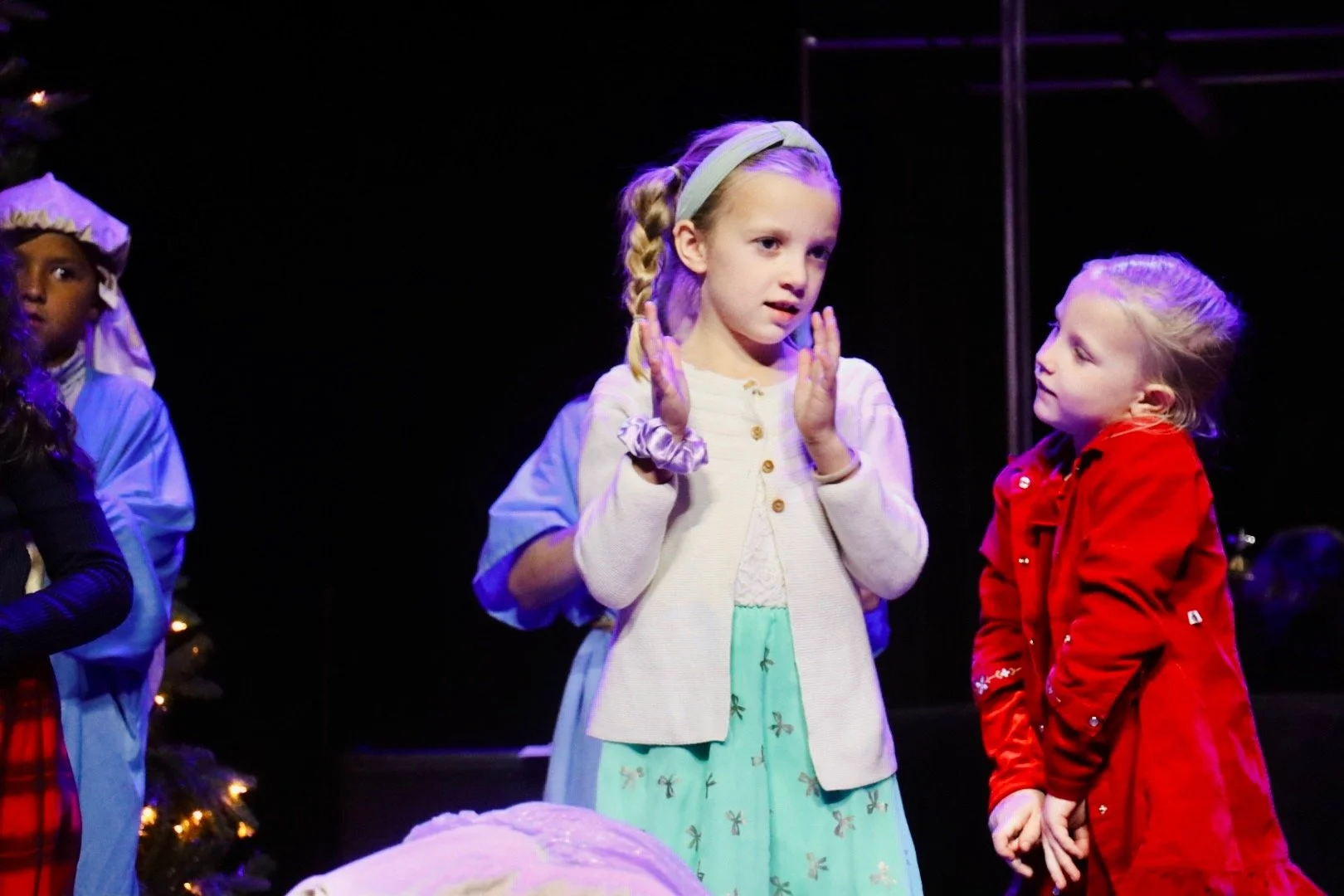 Children dressed in costumes performing on stage, with two girls in the foreground, one in a cream cardigan and mint dress, the other in a red jacket, engaged in a scene.