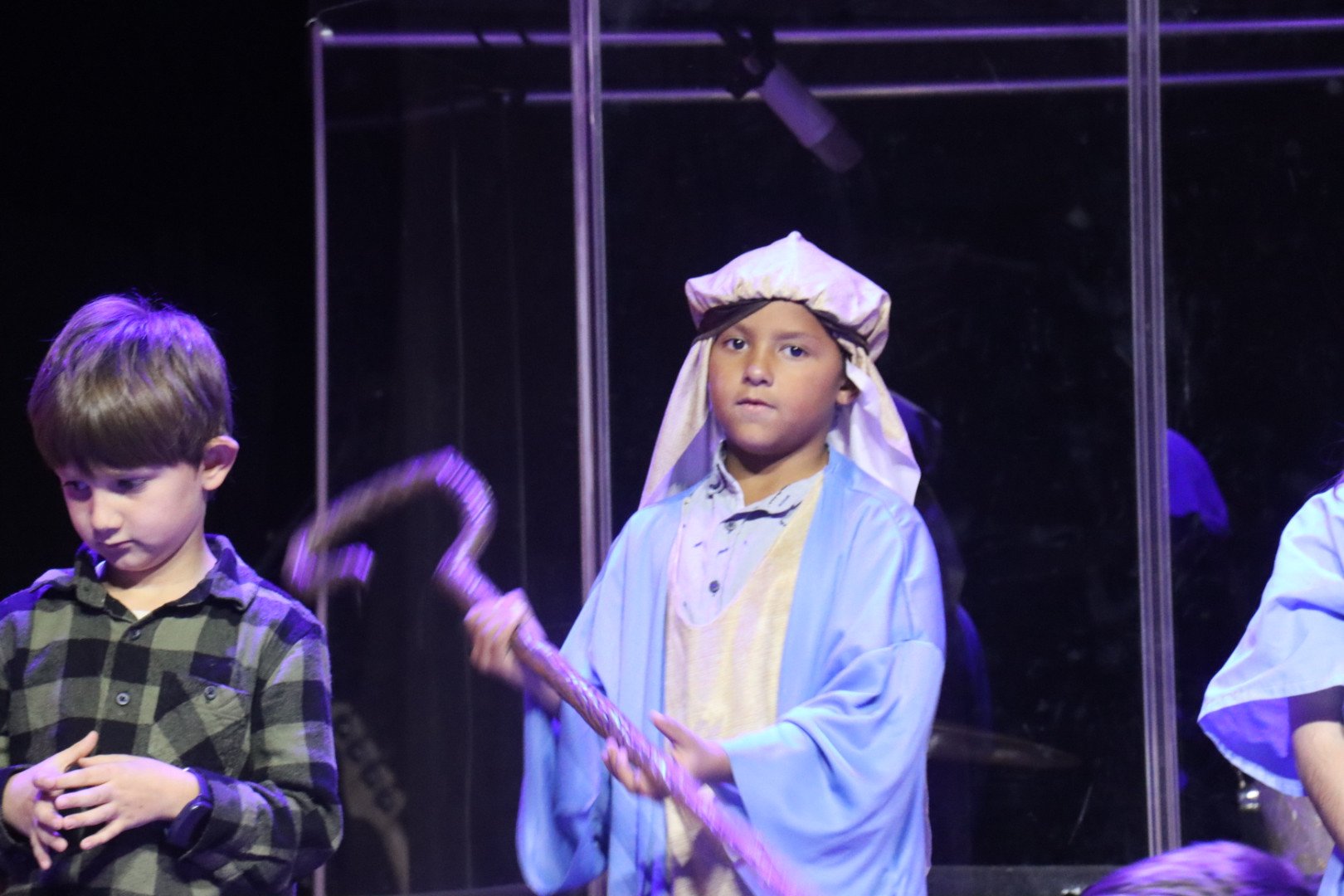 Children participating in a school play or nativity scene, with a boy dressed as a shepherd holding a staff, standing next to another boy in a plaid shirt, behind a glass or plastic barrier.