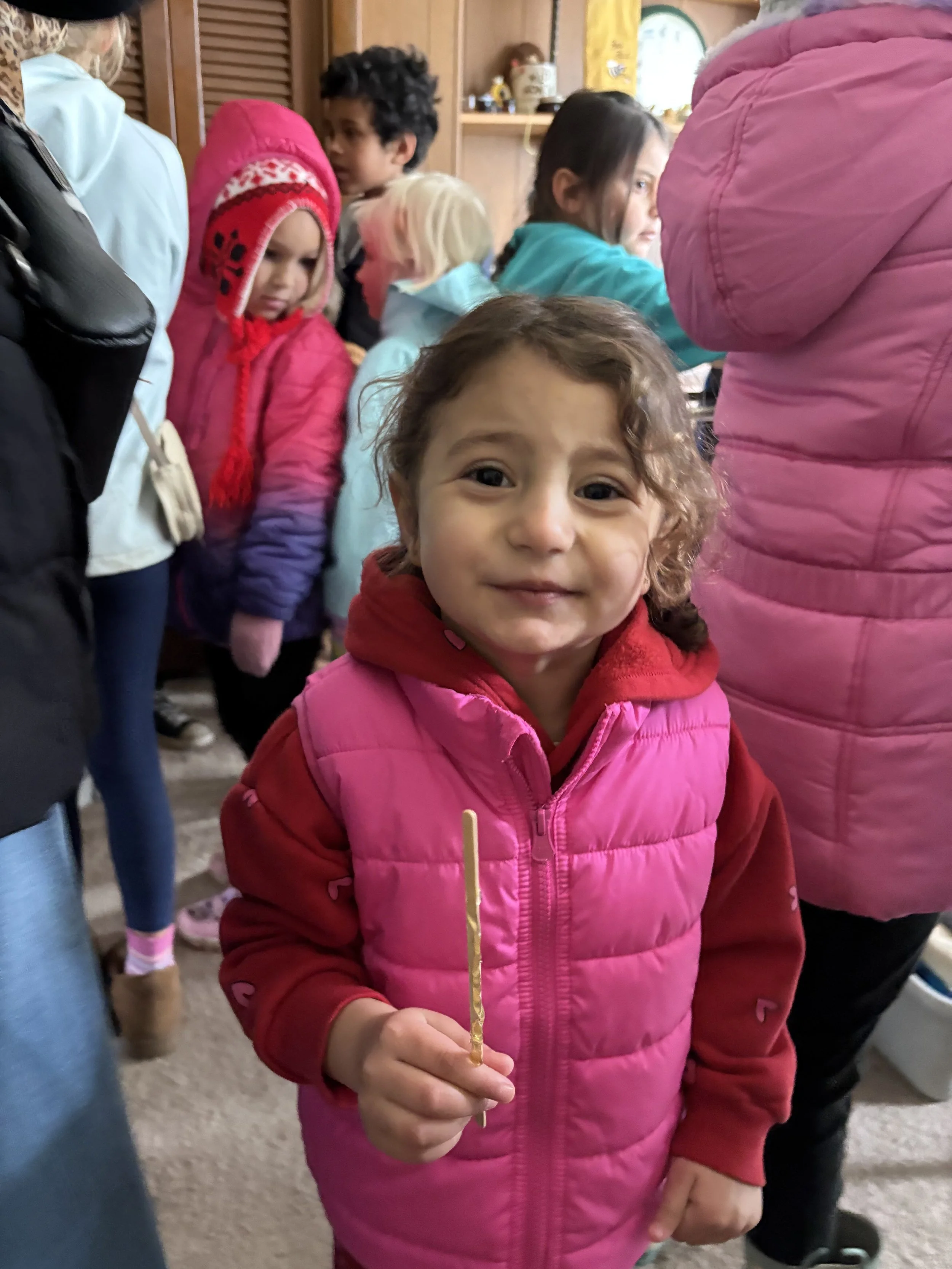A young girl with curly hair wearing a pink vest and red hoodie holding a yellow stick in a crowd of children and adults indoors.