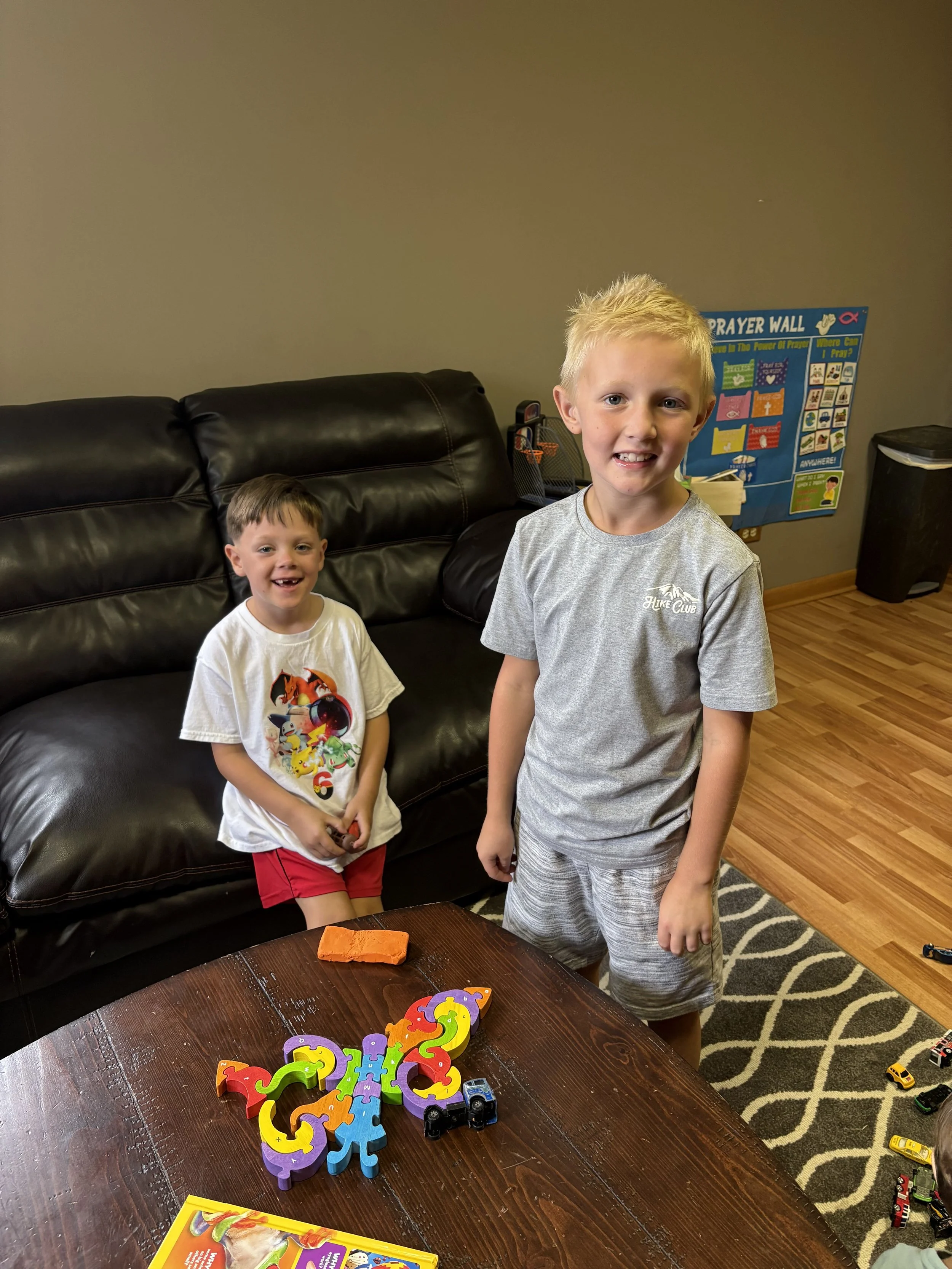 Two young boys playing with colorful number puzzle pieces and toy cars on a wooden table in a living room.