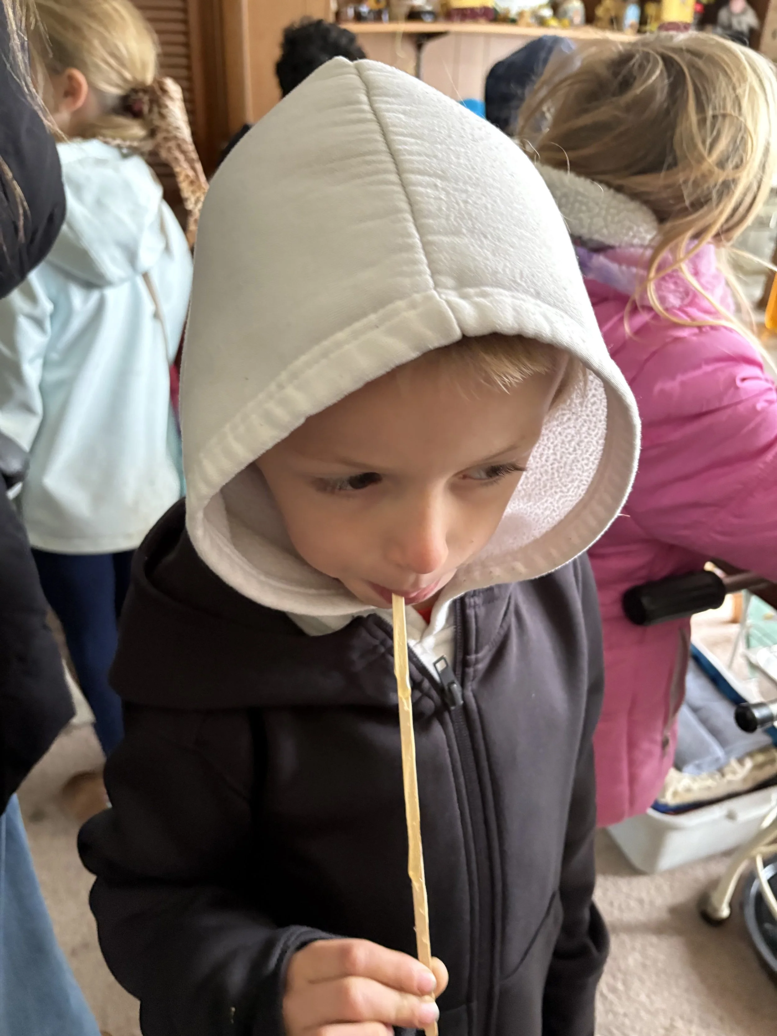 A young boy wearing a white hoodie and black jacket, holding a long, thin piece of caramel candy with a stick, licking it in a room with other children.