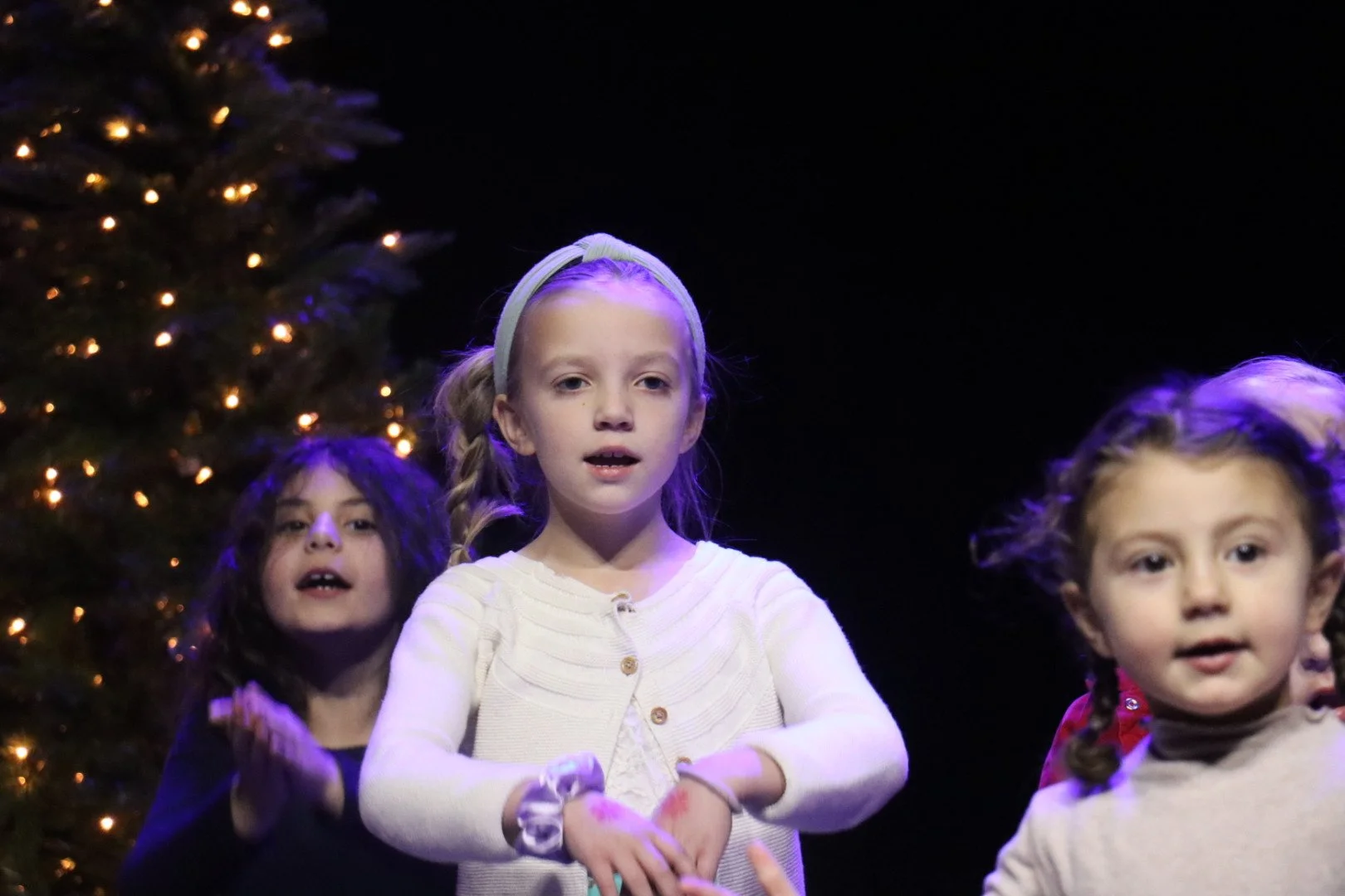 Three young girls singing or performing on stage with a decorated Christmas tree in the background.