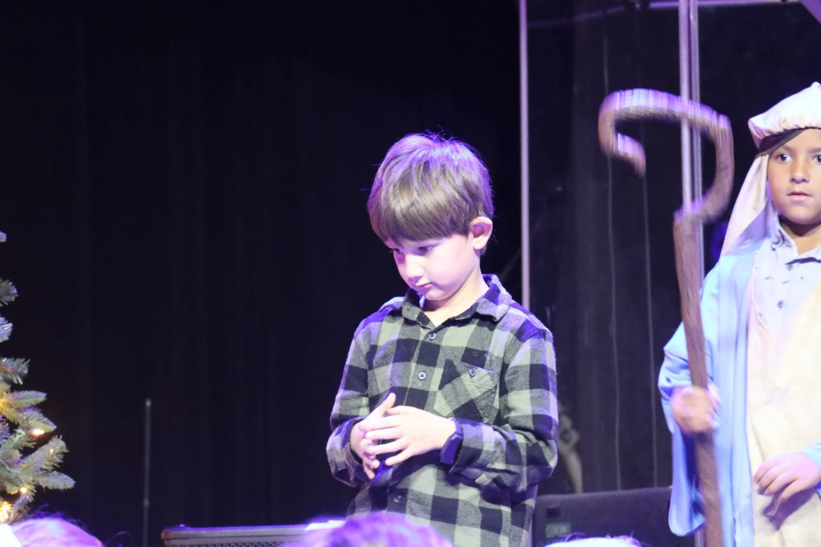 Two young boys participating in a school play. One is dressed as a shepherd holding a staff, the other in a plaid shirt with hands clasped, standing near a decorated Christmas tree on stage.