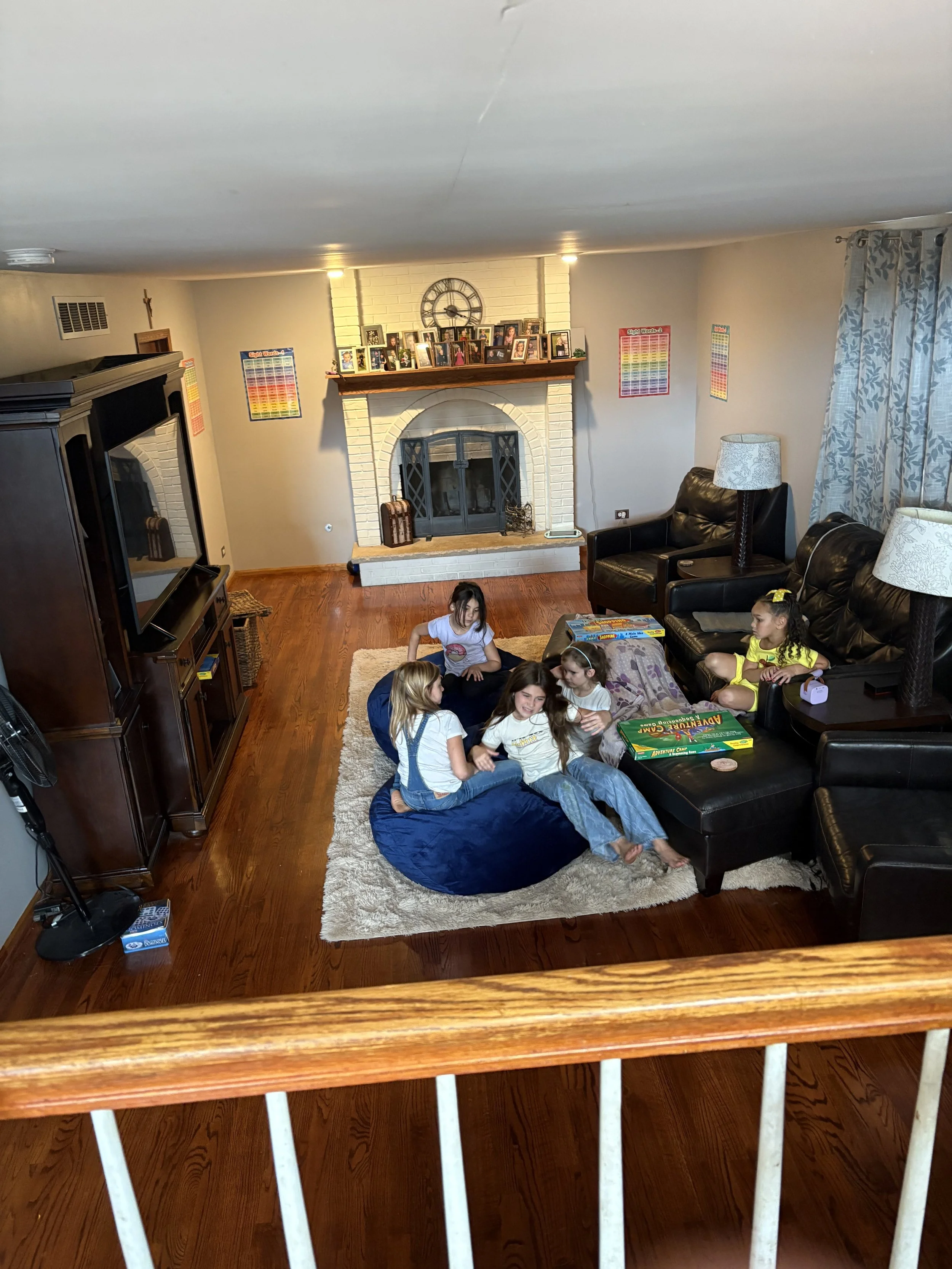 A family gathering in a living room with five children ages 4-10, seated on a cream-colored rug and blue bean bag chair, playing a board game together. The room features a brick fireplace with family photos on the mantel, a large TV, leather couches,