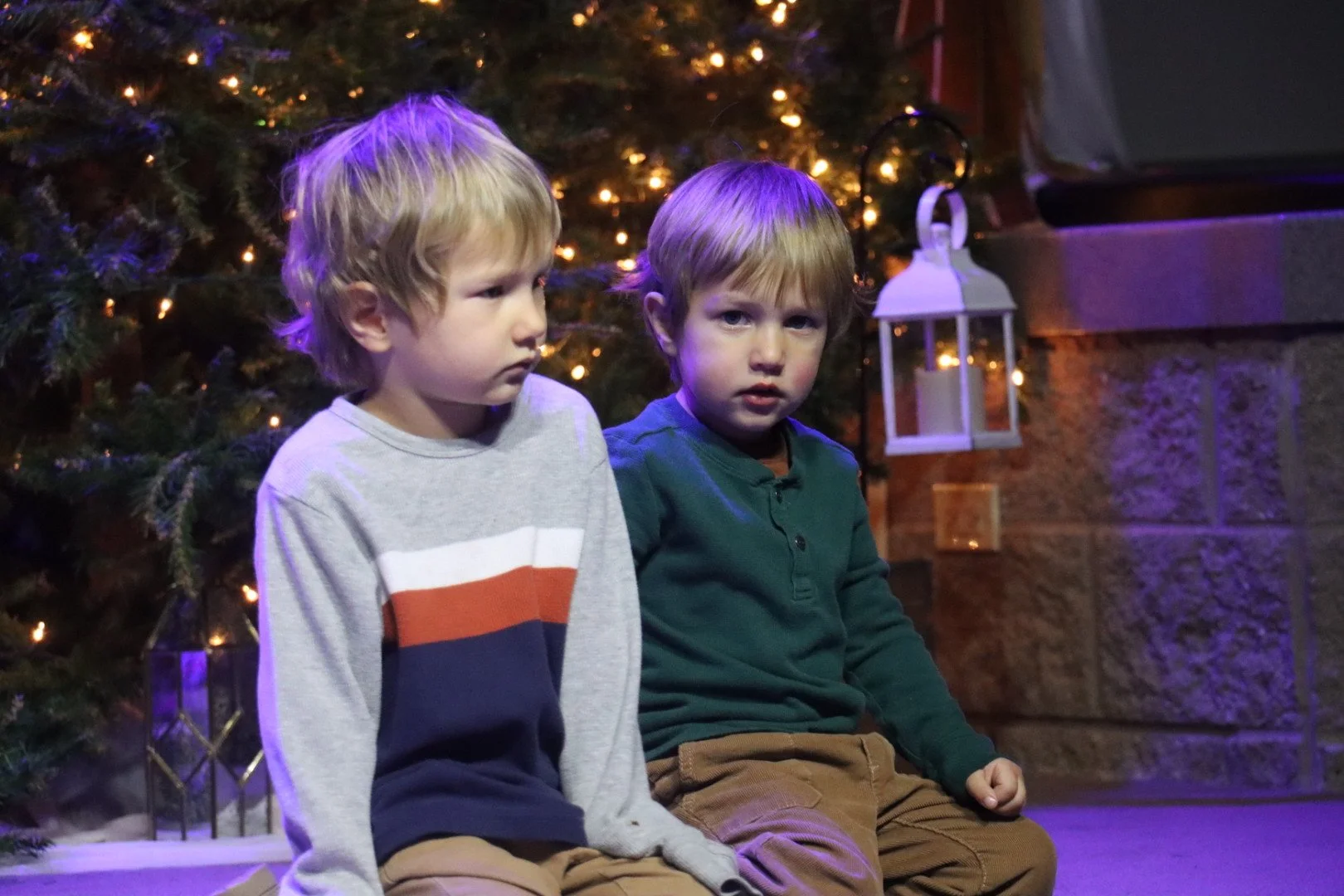 Two young boys with light brown hair sitting in front of a decorated Christmas tree with lights, near a wall with a lantern hanging.