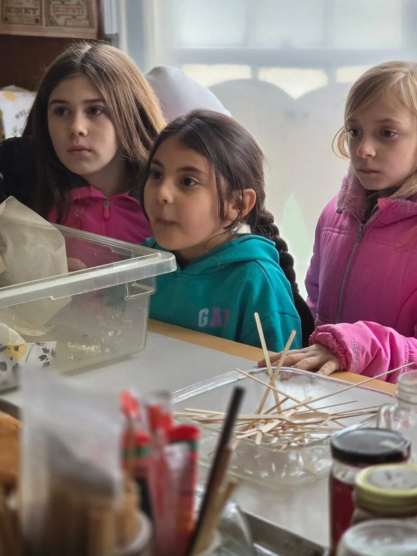 Three young girls observing a baking or craft project on a table, with various materials like straws and food items nearby.