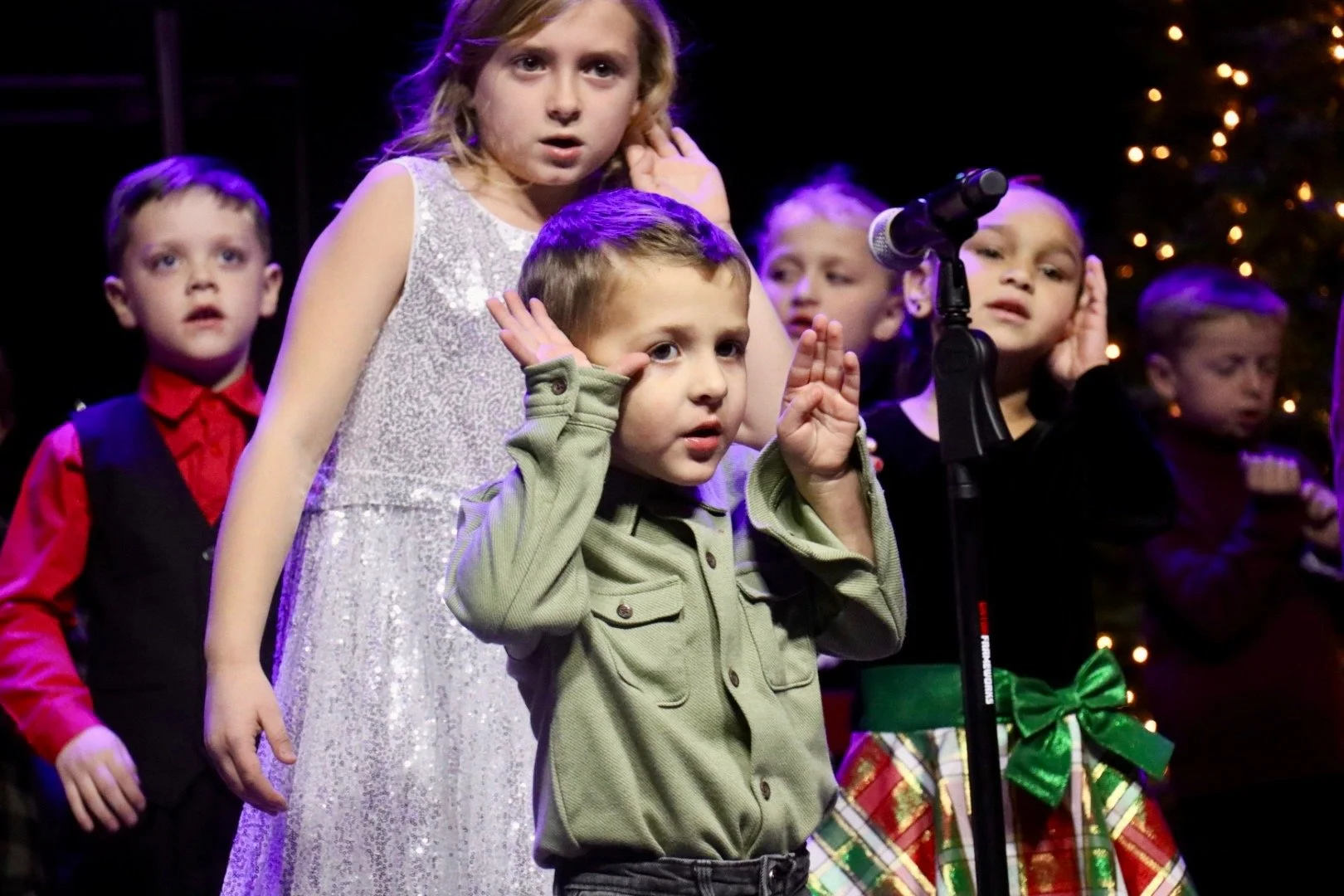 Children performing on stage during a holiday event, with a Christmas tree and lights in the background. One boy at a microphone appears to be singing or speaking, while others wait or perform behind him.