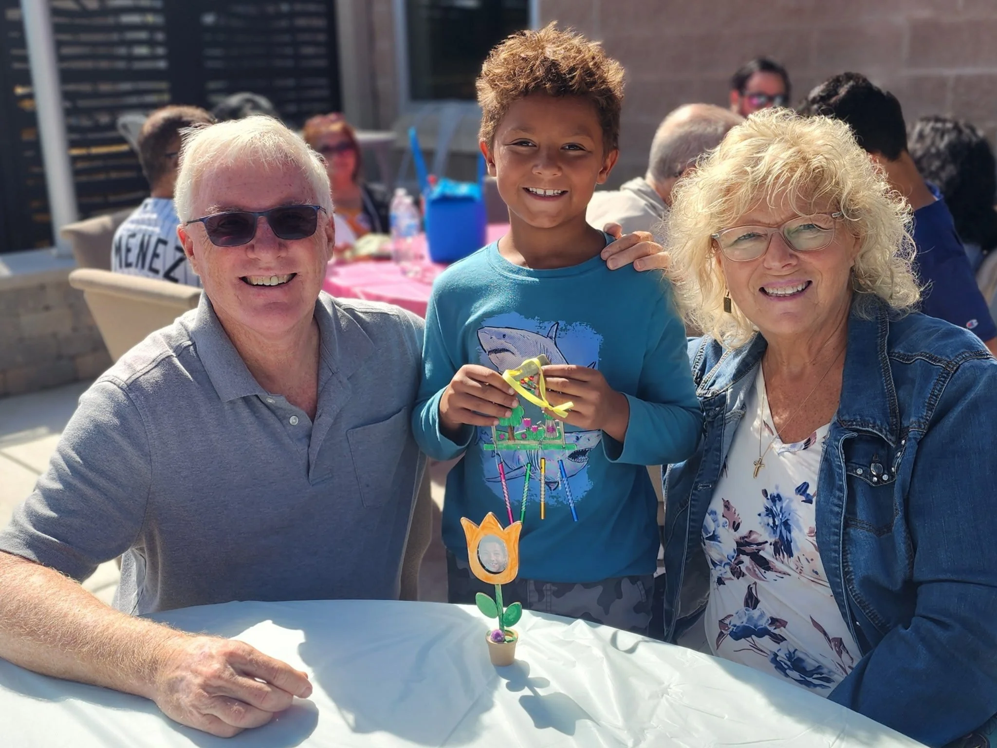 Three people smiling at an outdoor celebration, with a woman, a teenage boy, and an older man, sitting around a table with a colorful handmade craft in front of them.