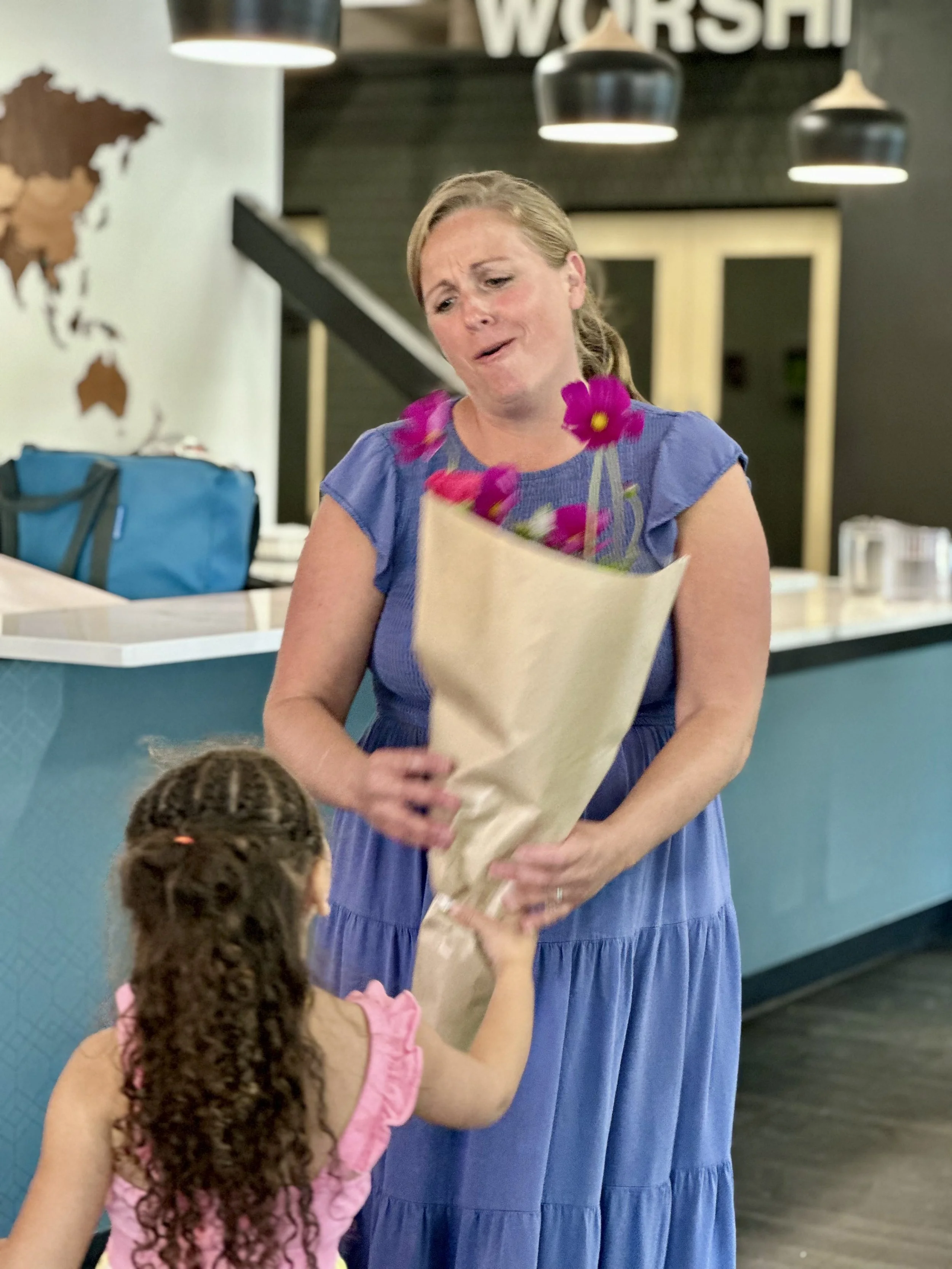 A woman in a blue dress accepting a bouquet of flowers from a young girl with curly hair in a pink dress inside a modern building.