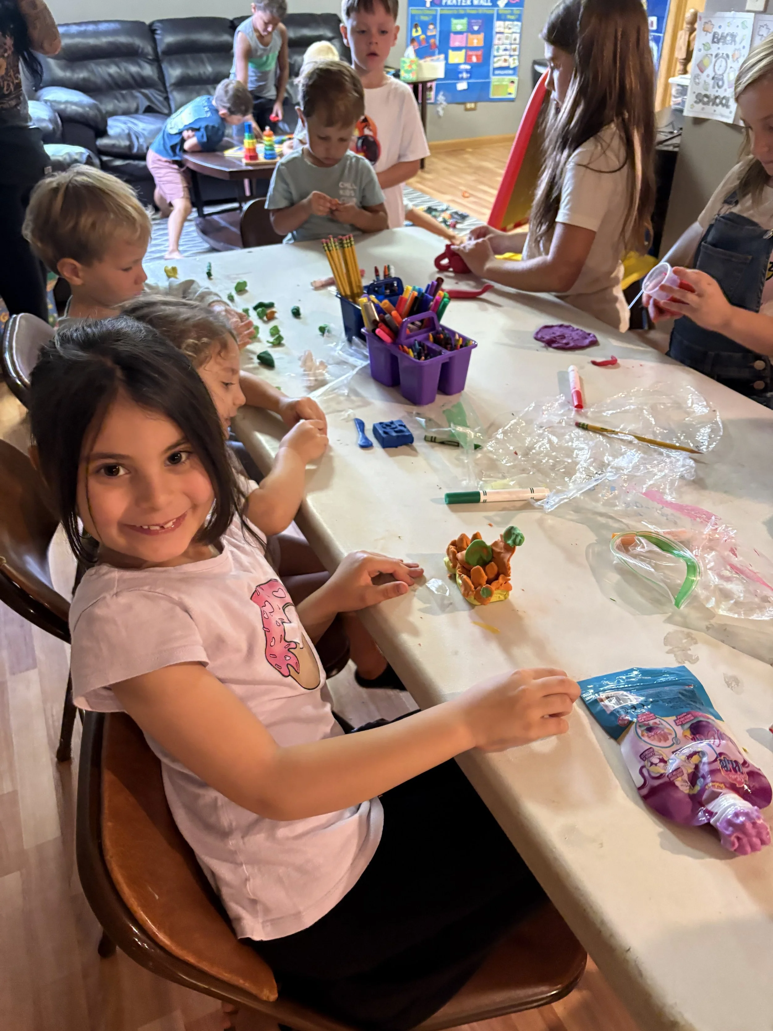 A group of children gathered around a table engaging in arts and crafts activities, with various craft supplies such as markers, clay, and other materials on the table.