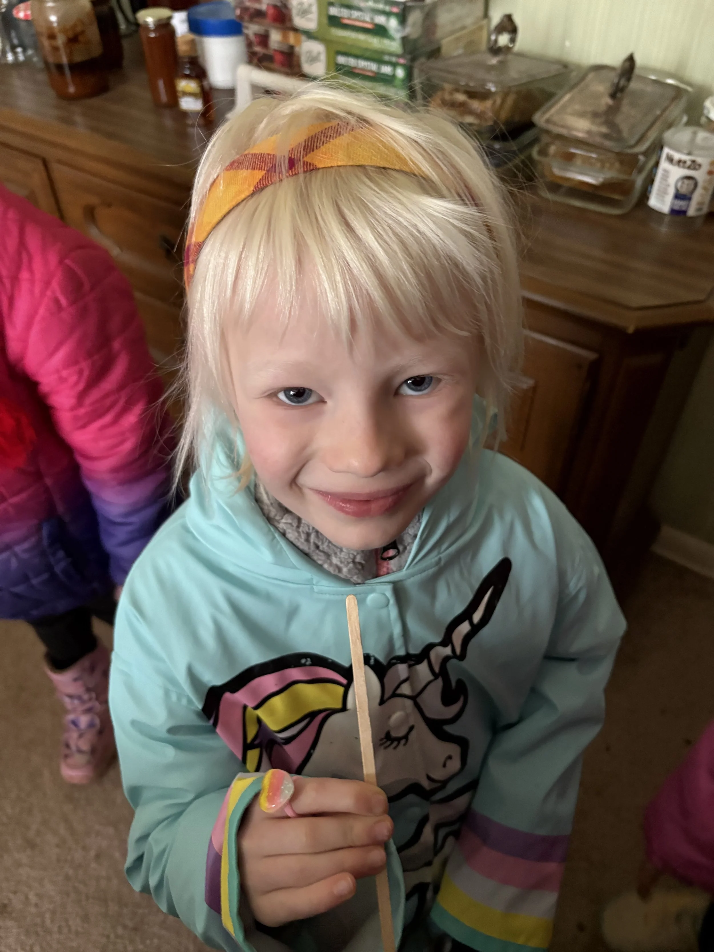 A young girl with blonde hair, blue eyes, and a colorful headband, smiling and holding a wooden stick inside a room with wooden cabinets and food containers.