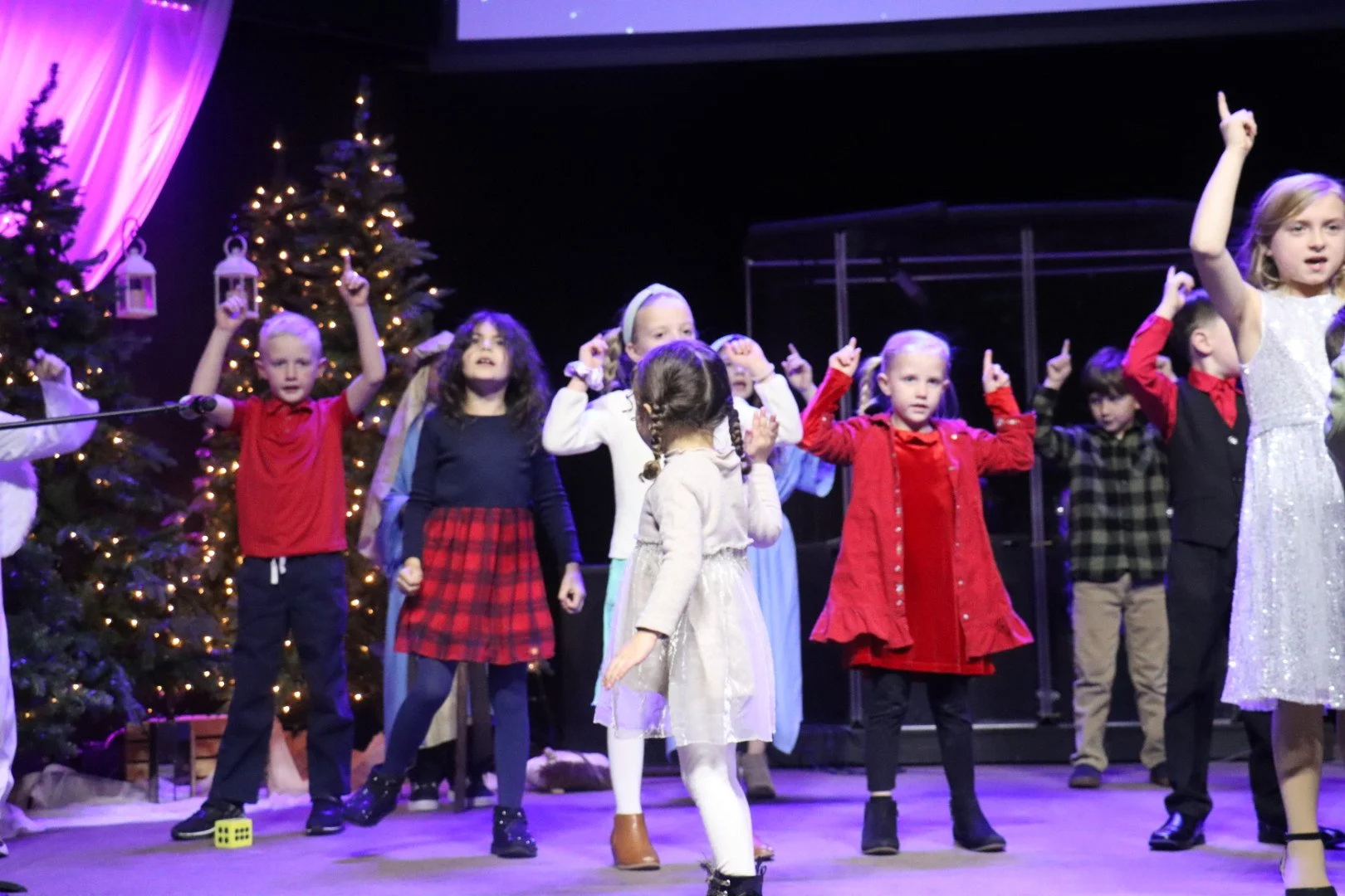 Children performing on stage during a holiday play, with Christmas trees decorated with lights in the background.