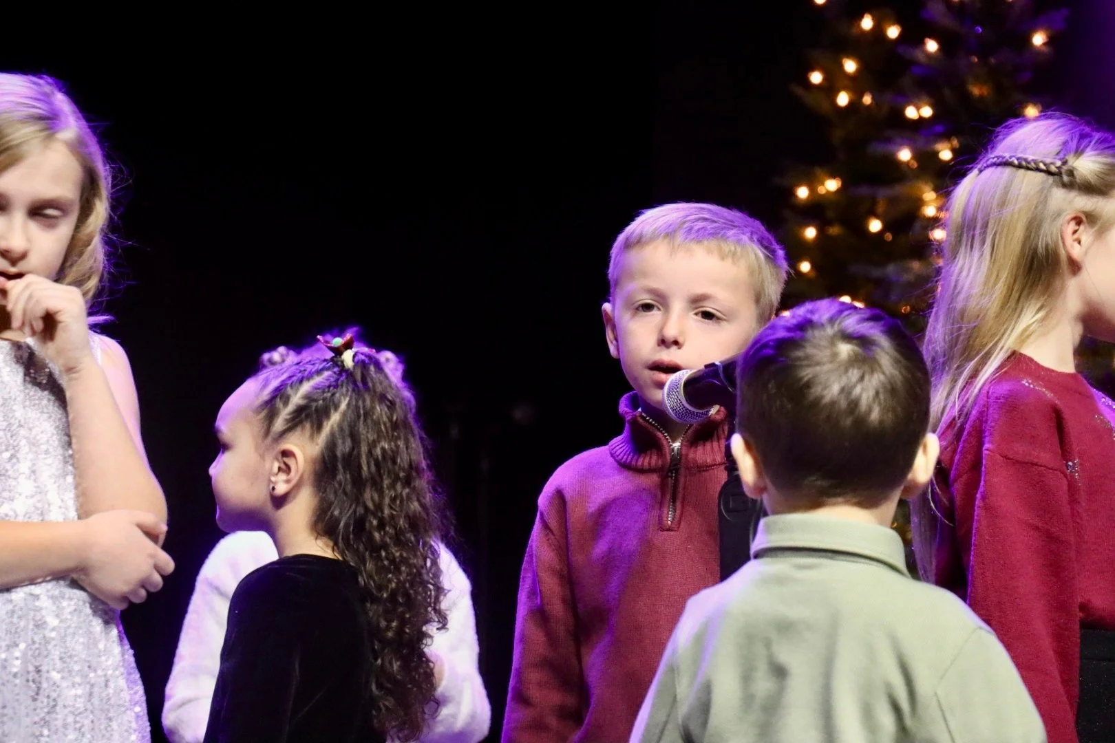 Children on stage during a Christmas performance, with a Christmas tree in the background.