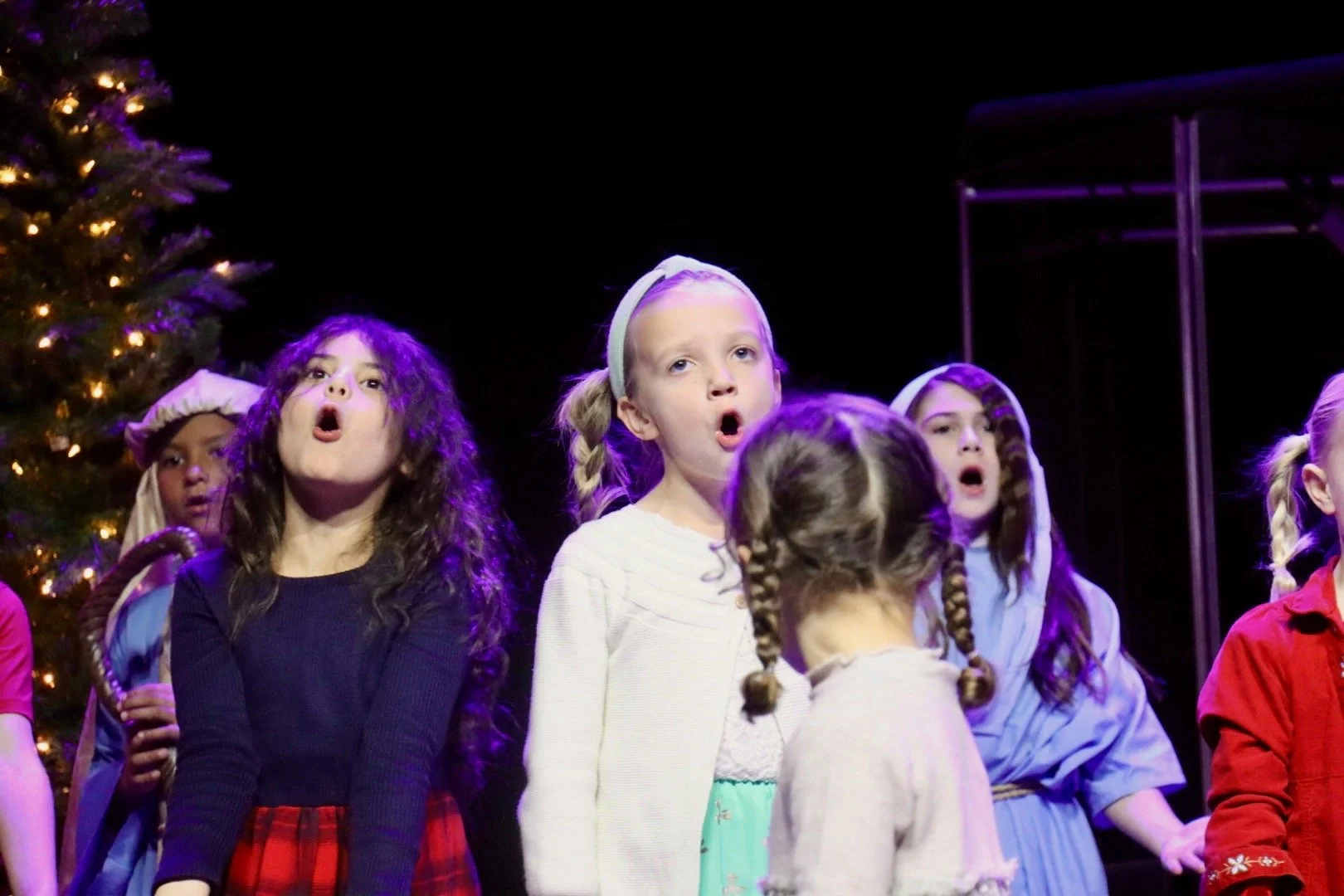 Children performing in a Christmas play, dressed in costumes, with a decorated Christmas tree in the background.