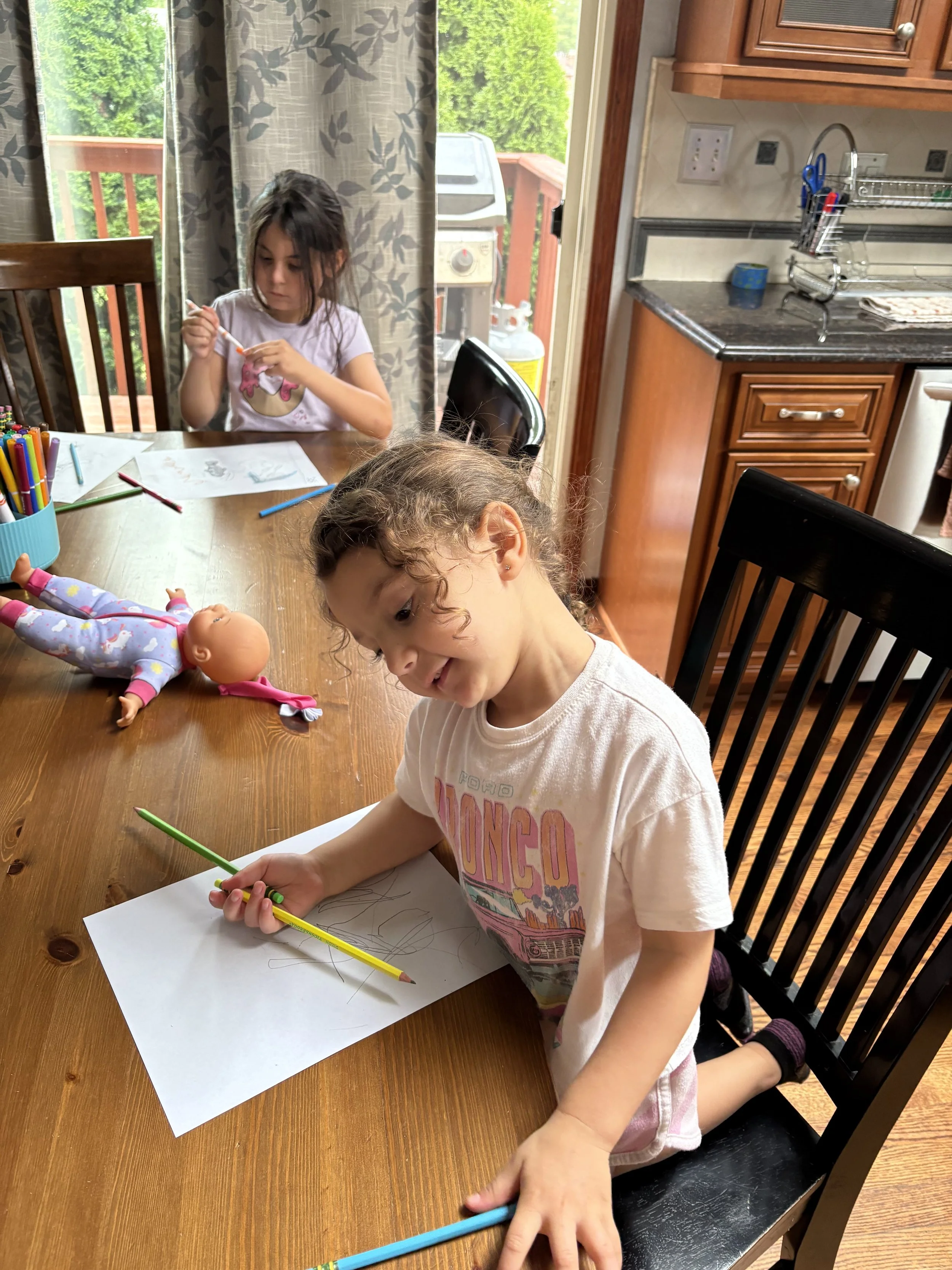 Two young girls at a wooden dining table engaged in drawing and coloring activities. One girl is smiling and holding a green-colored pencil, while the other girl is sitting at the far end looking at her craft. The table has colorful markers, pens, an