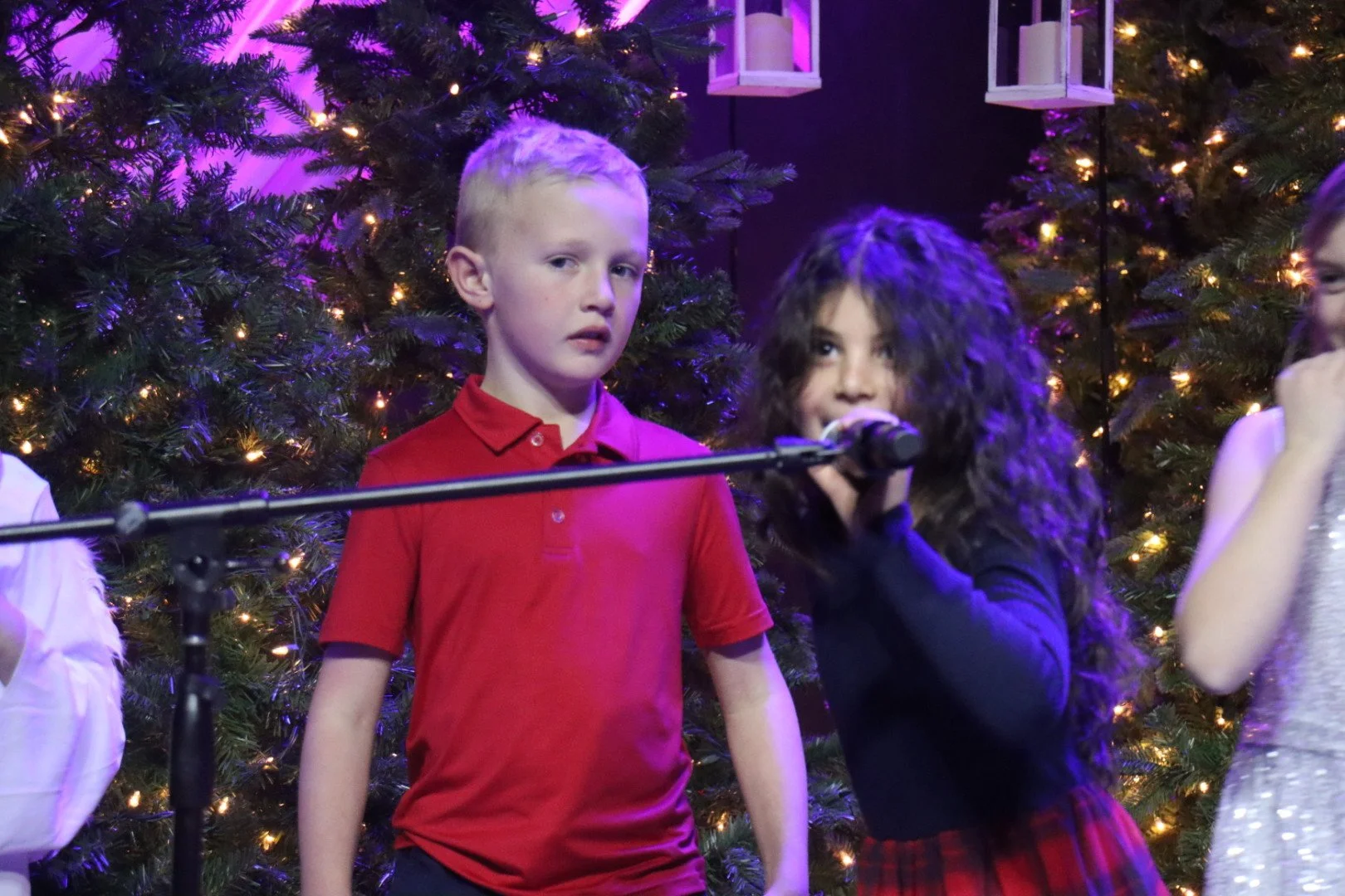 Children singing or performing on stage in front of a decorated Christmas tree with lights.