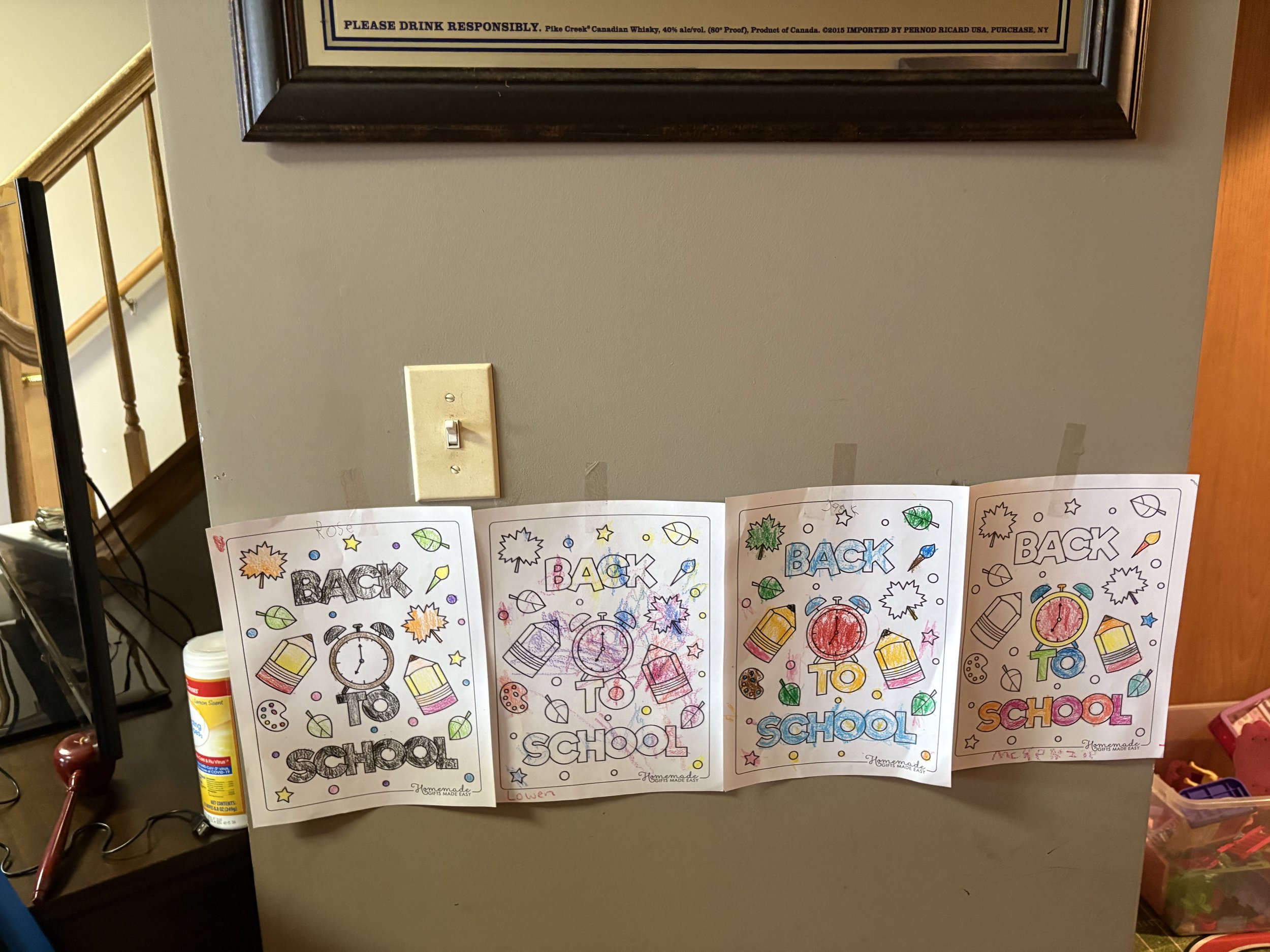 Four colorful back-to-school coloring pages taped to a wall, each reading 'Back to School' with various school-related drawings. The pages are decorated with crayons, books, clocks, and leaves, and feature different handwriting and signatures at the 