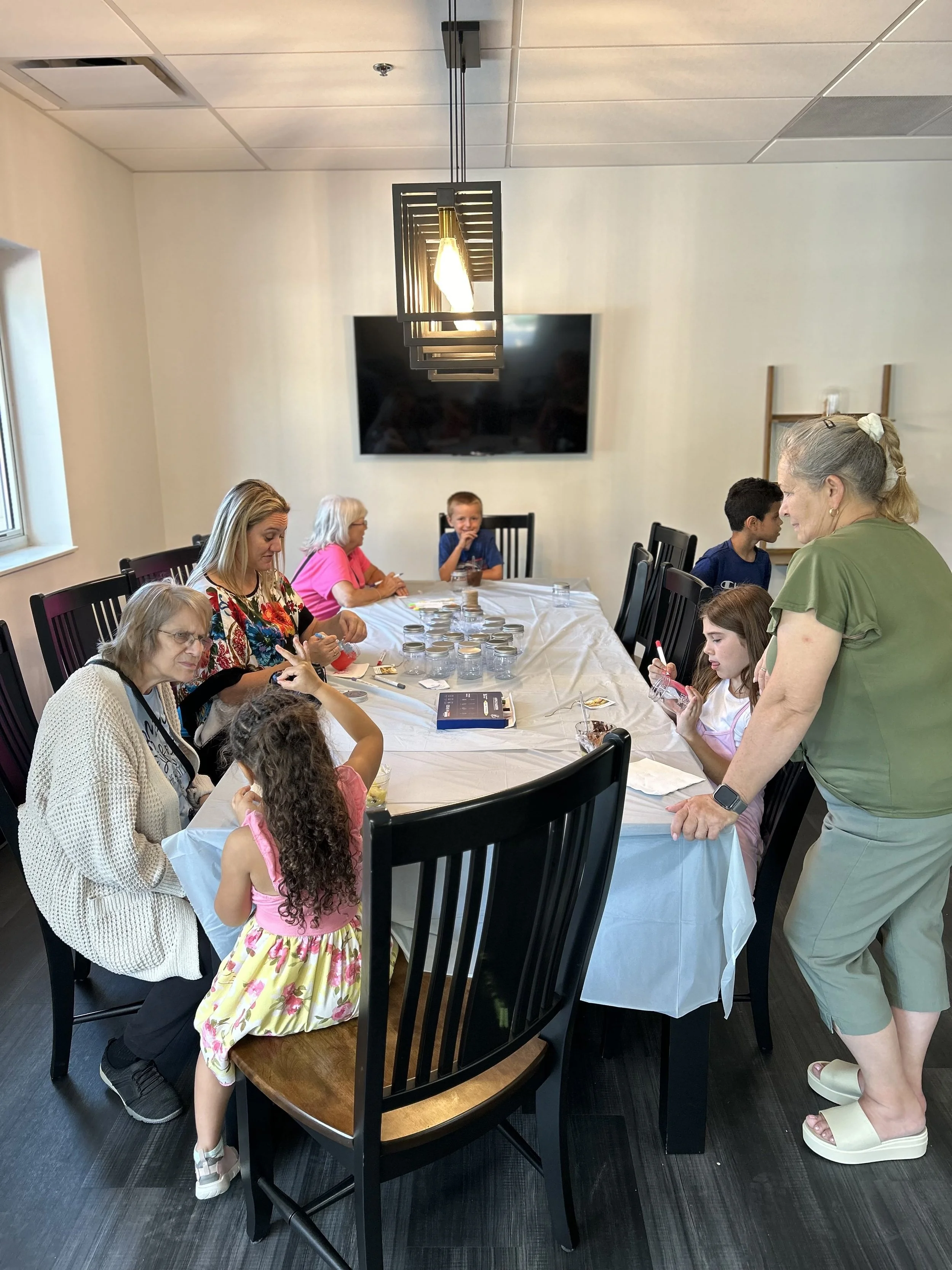 A group of people, including children and adults, gathered around a dining table in a room, engaging in a hands-on activity or workshop. There are jars and materials on the table, and one woman is assisting a girl who is eating or making an item. The