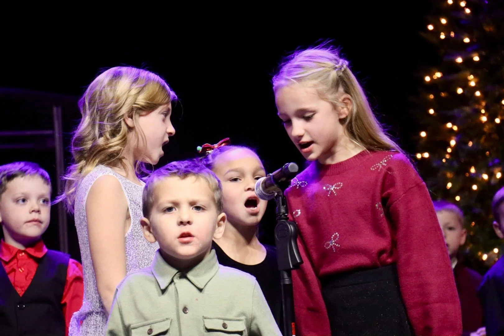 Children singing on stage during a Christmas performance, with a Christmas tree decorated with lights in the background.