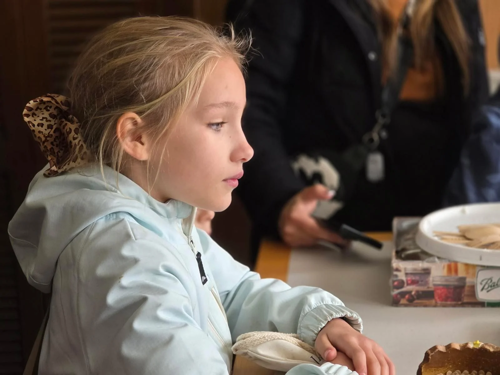 A young girl with blonde hair tied with a leopard print bow, sitting at a table with her arms crossed, wearing a light-colored jacket, and looking to her left in a cozy indoor setting.
