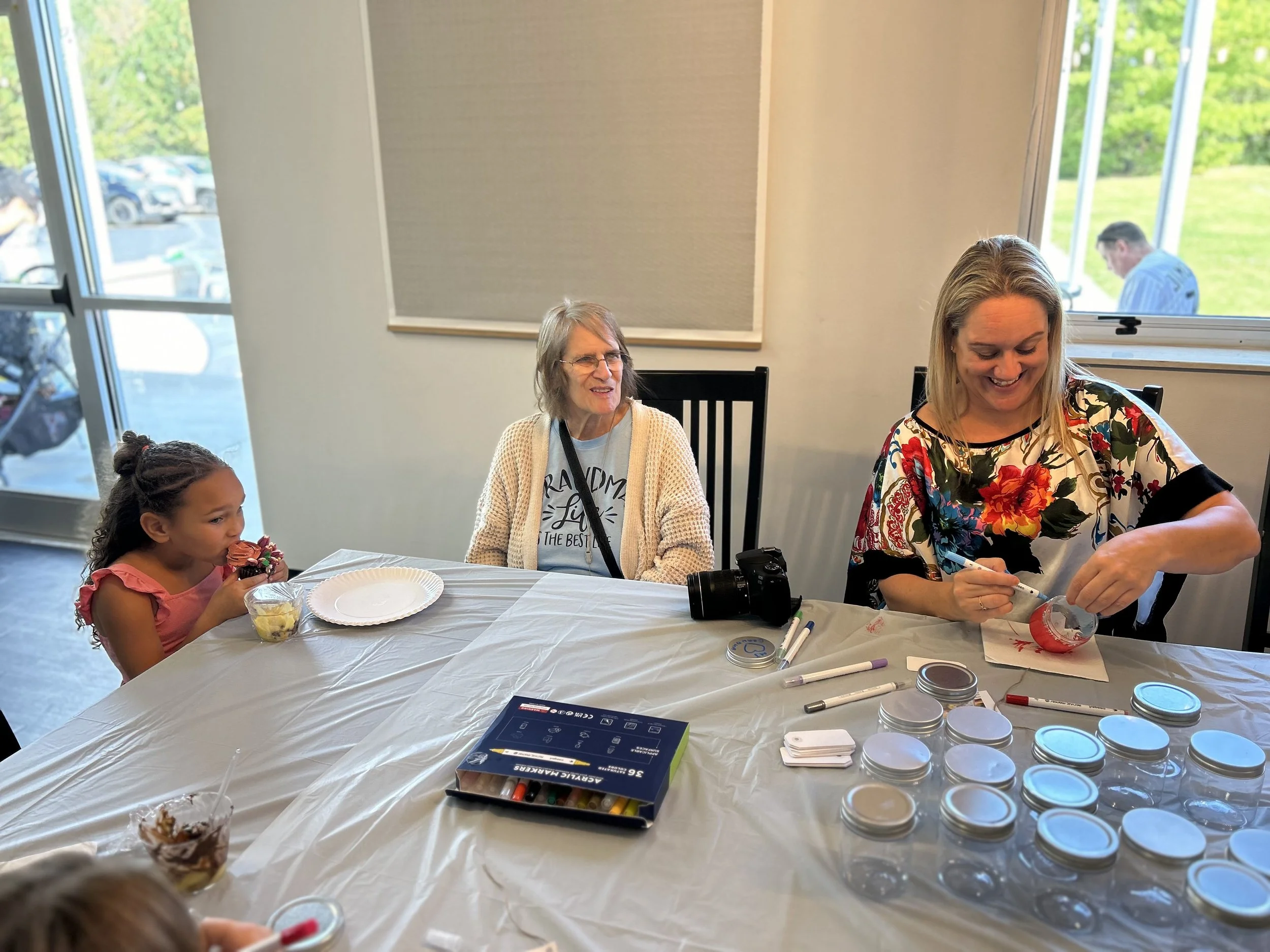 A woman decorating jars with red paint while sitting at a table with two other women and a young girl with ice cream. There are jars, markers, and a camera on the table.