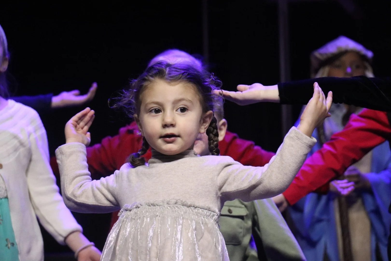 A young girl with braided hair and a beige dress performs on stage with other children, gesturing with her hands during a performance.