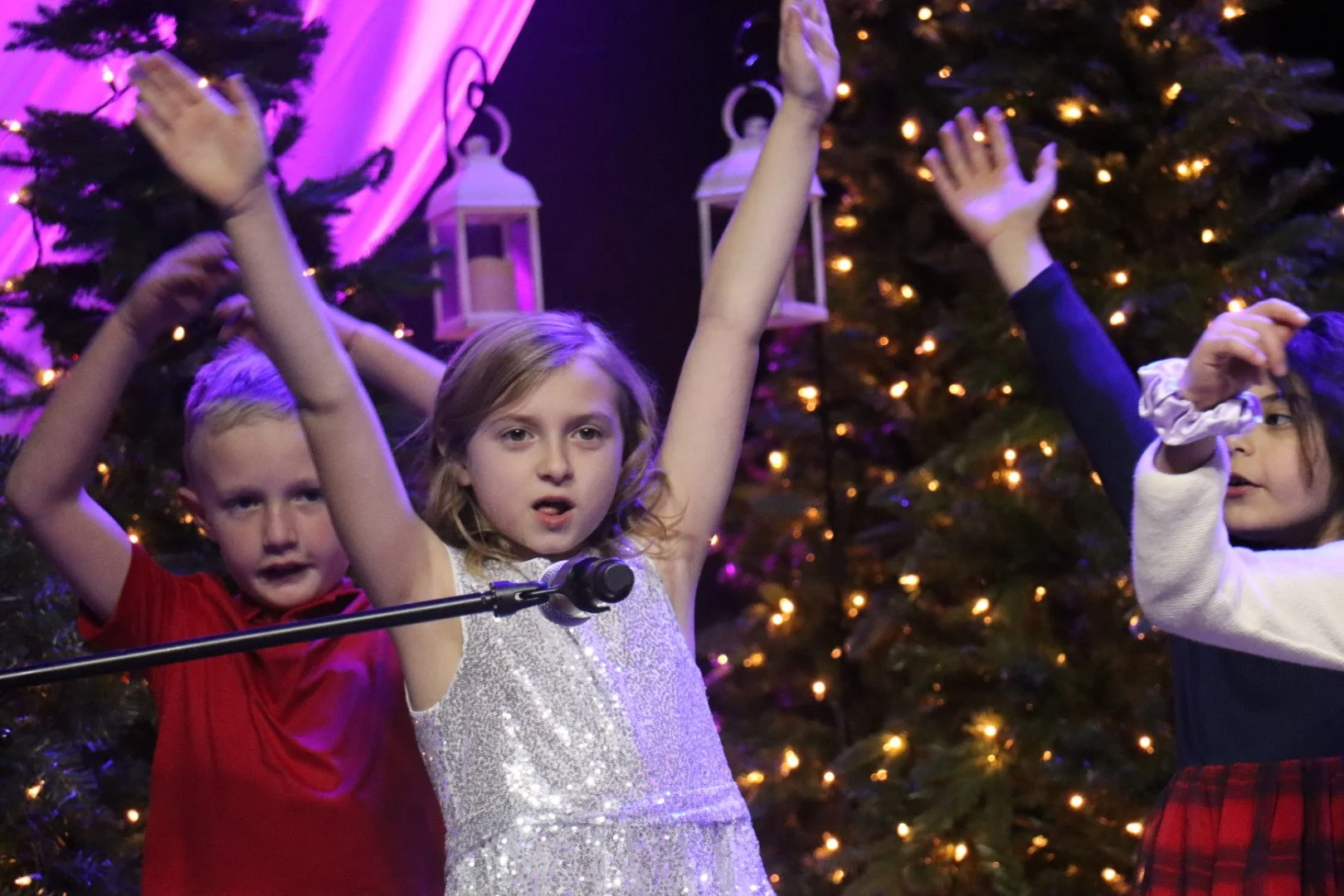 Three children performing on stage with Christmas trees and lights in the background.