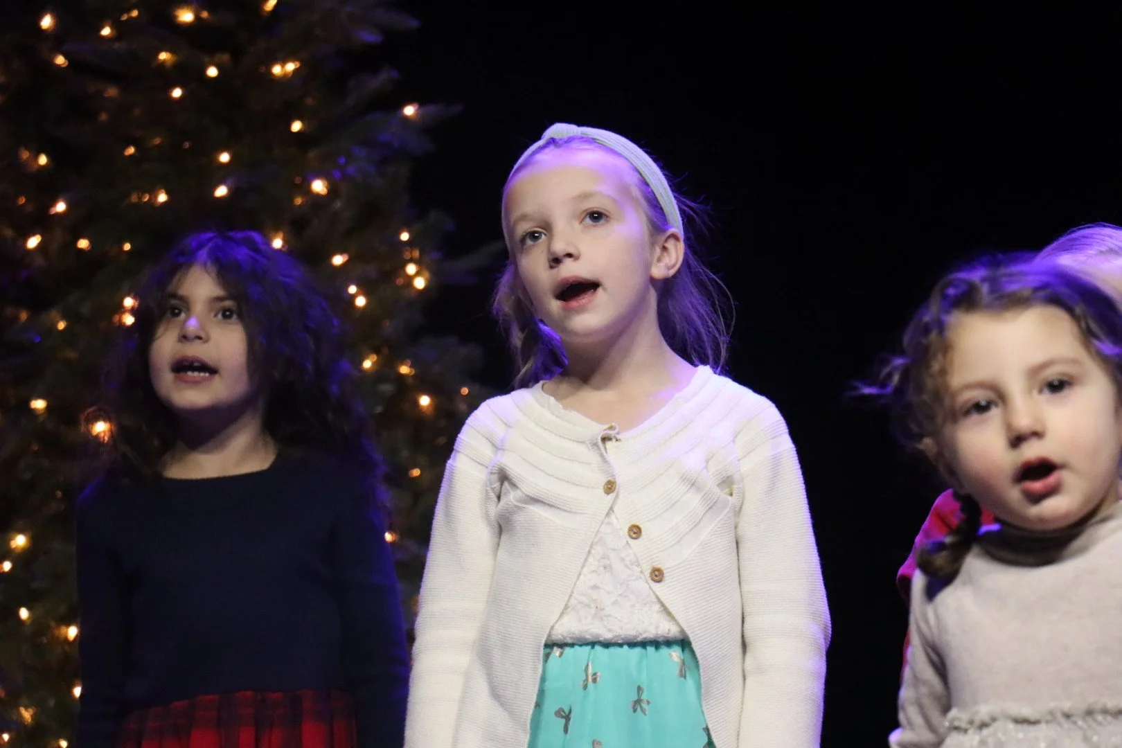 Three young girls singing or speaking on stage in front of a decorated Christmas tree with lights.