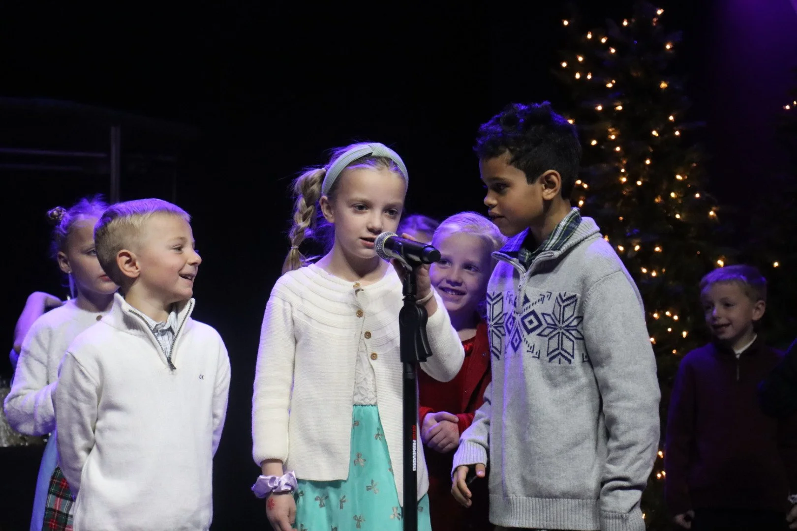 Children on stage during a holiday performance, with a Christmas tree in the background, one girl speaking into a microphone, surrounded by other children in festive clothing.