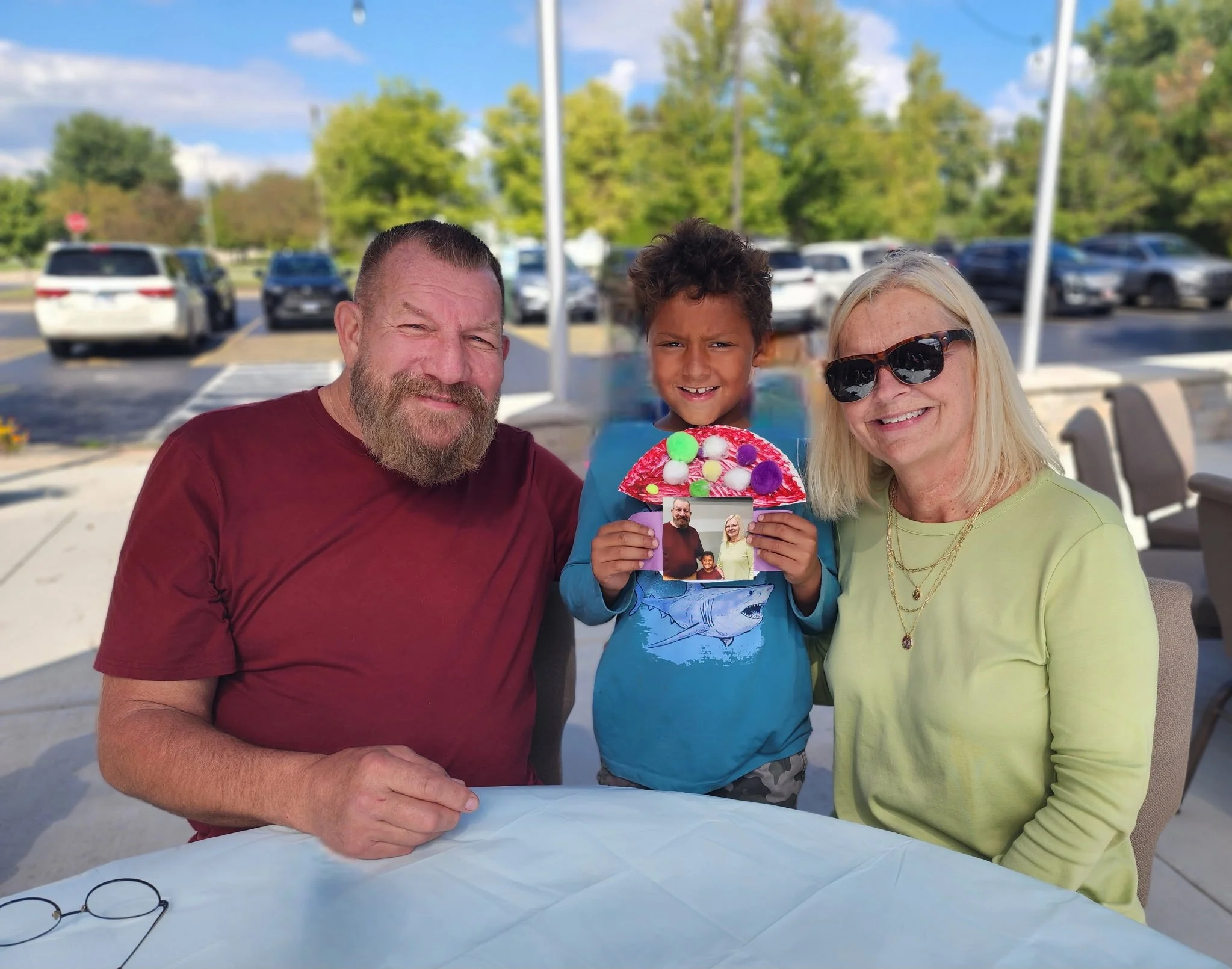 A family of three sitting at a table outdoors with parked cars and trees in the background. The child is holding a colorful, handmade craft and a small photograph.