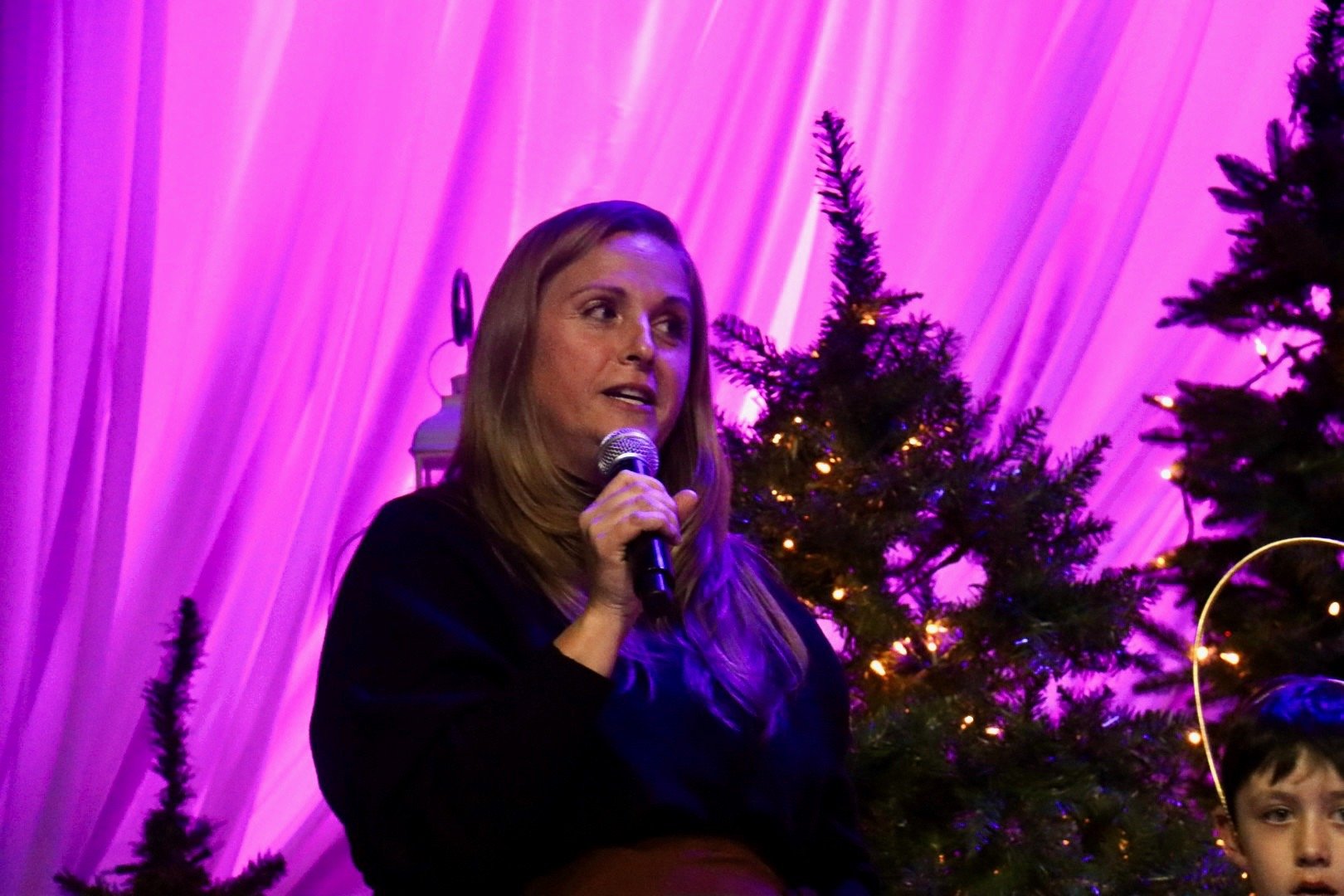 A woman with long brown hair speaking into a microphone, sitting near decorated Christmas trees with lights, in front of purple draped backdrop.