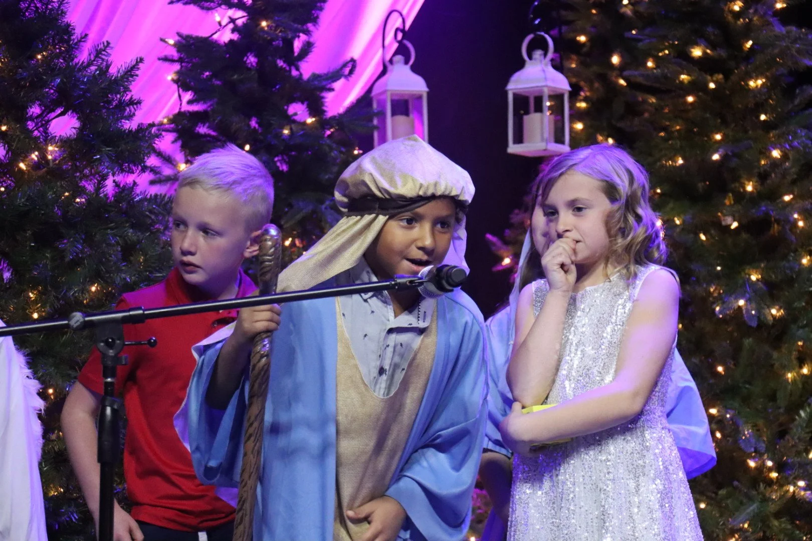 Children dressed as nativity characters performing in a Christmas play with decorated Christmas trees and lanterns in the background.