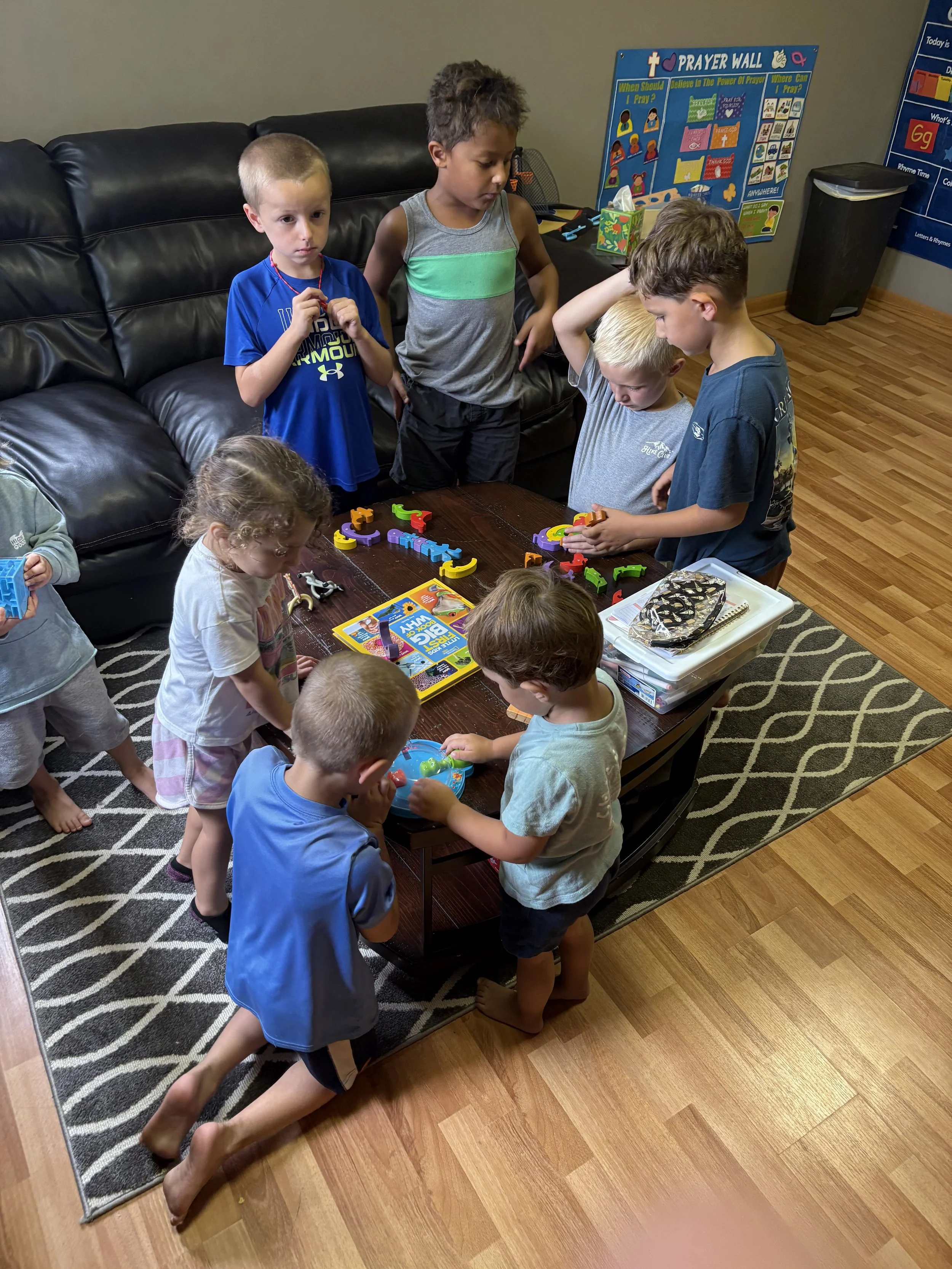 Group of children gathered around a table playing with toys in a room with educational posters on the wall.