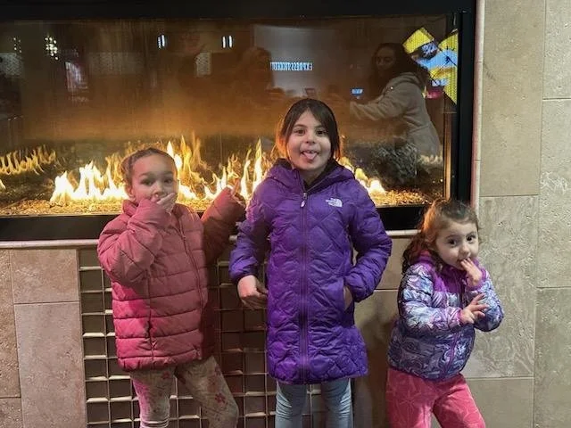 Three young girls in colorful winter jackets standing in front of a fireplace with flames, smiling and posing for the photo.
