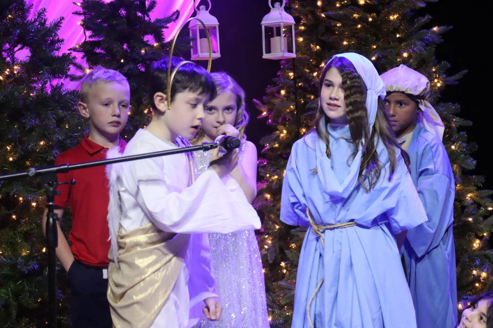 Children dressed in costumes performing a Christmas play on stage decorated with Christmas trees and lanterns.