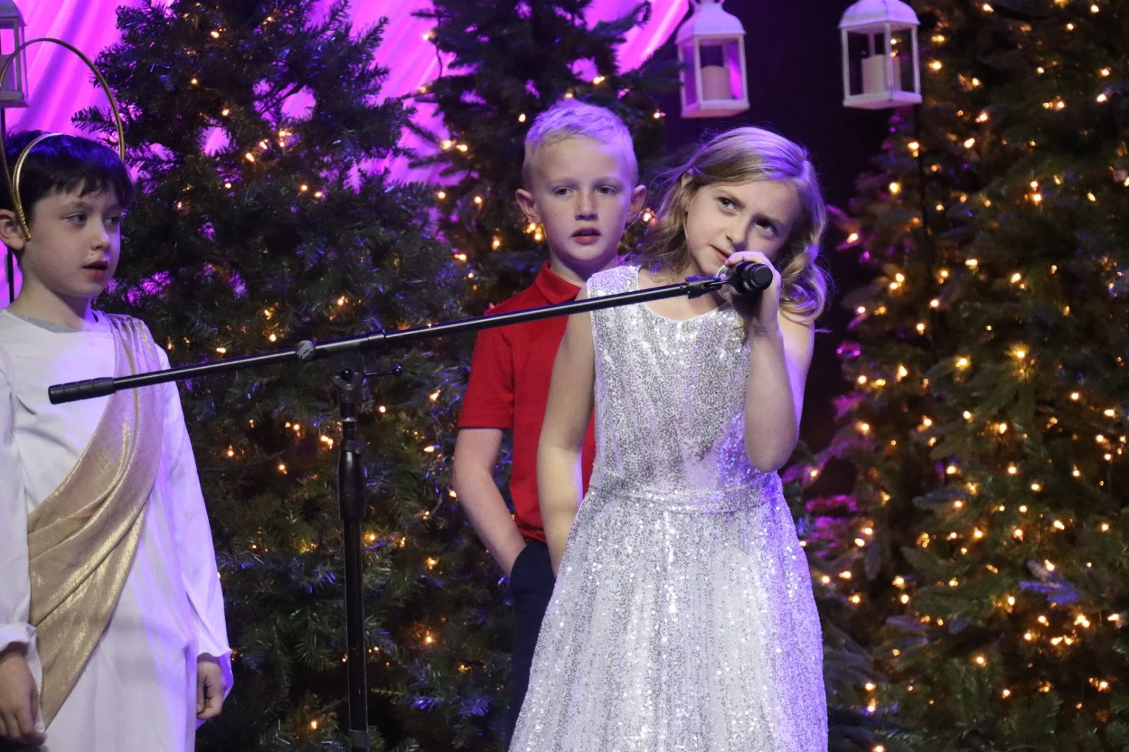 Children performing on stage during a Christmas concert, with decorated Christmas trees and lanterns in the background.