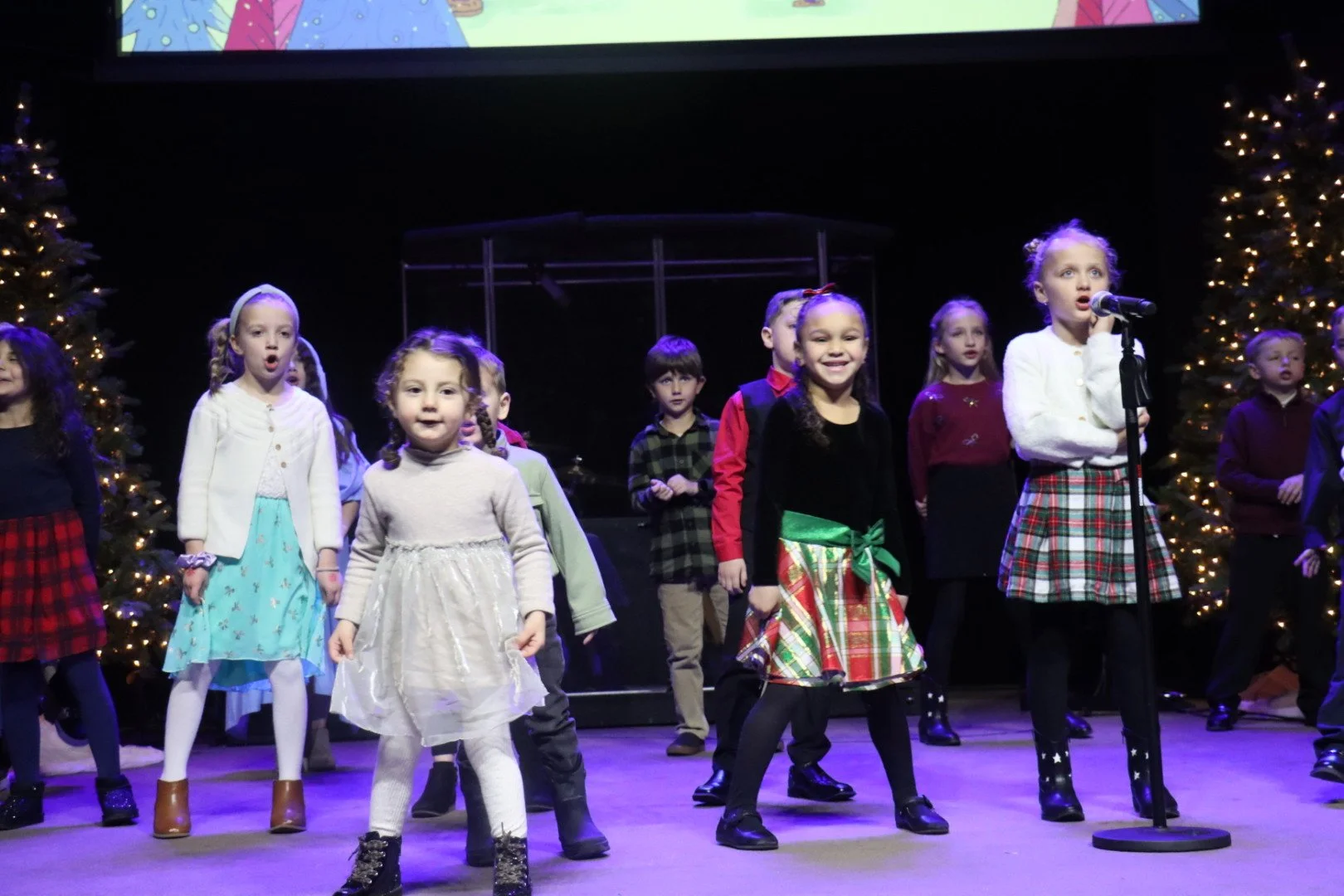 Children performing in a Christmas school play, dressed in festive clothing, on stage decorated with Christmas trees and lights.