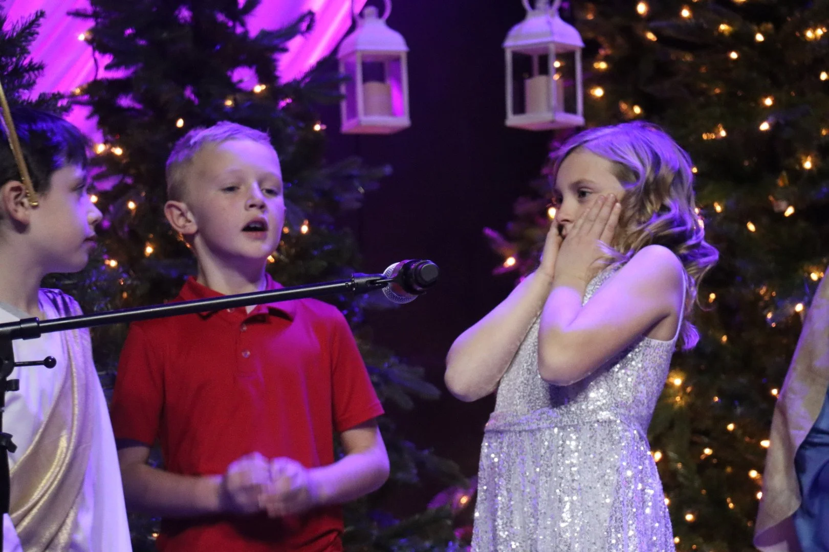 Three children standing in front of a decorated Christmas tree during a holiday performance; one girl with curly hair is surprised, a boy in a red shirt appears to be speaking or singing, another girl on the left is listening.