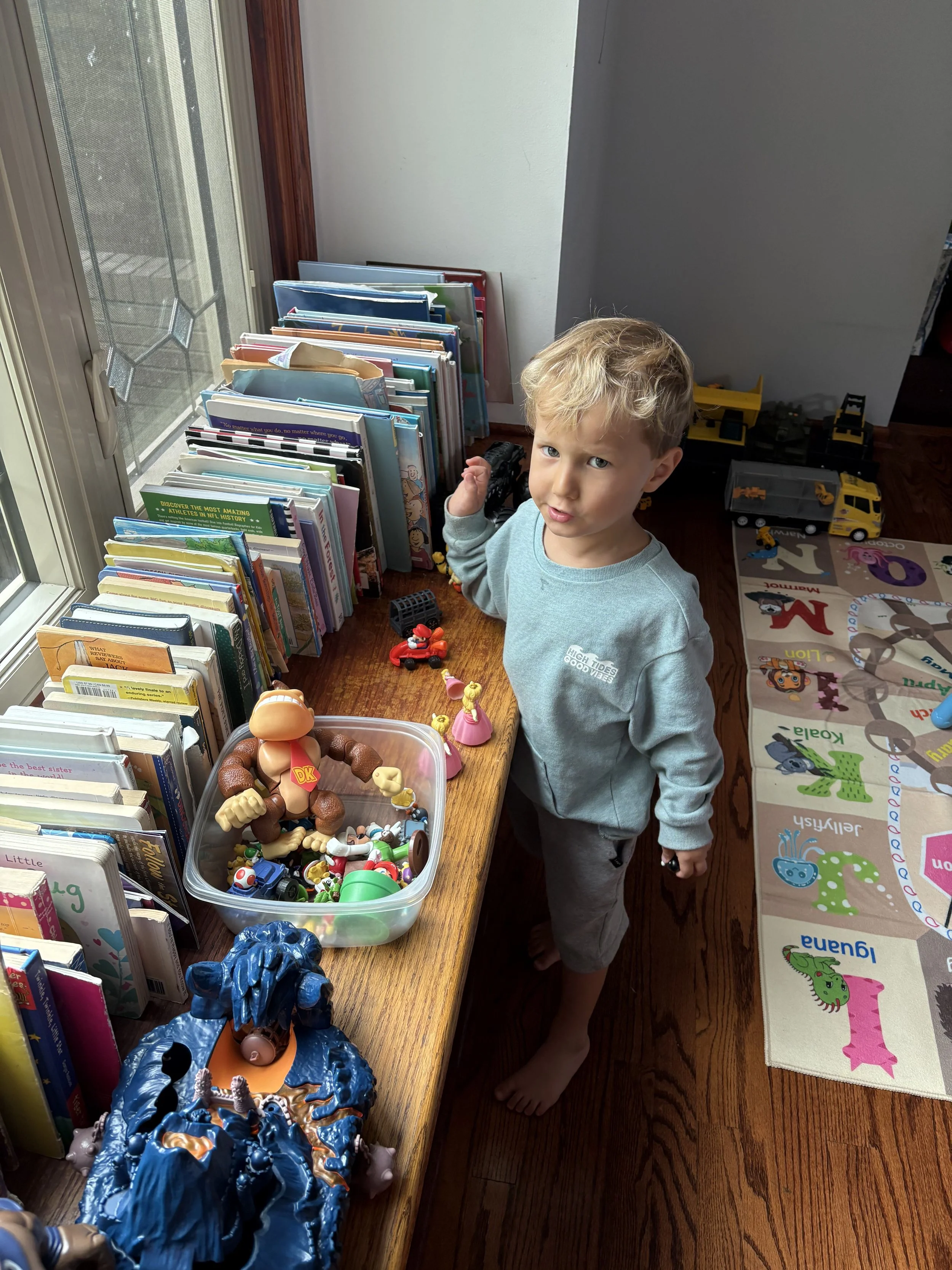 A young boy with blonde hair standing barefoot next to a wooden table filled with books and toys, near a window with a screened door, in a room with an alphabet-themed rug.