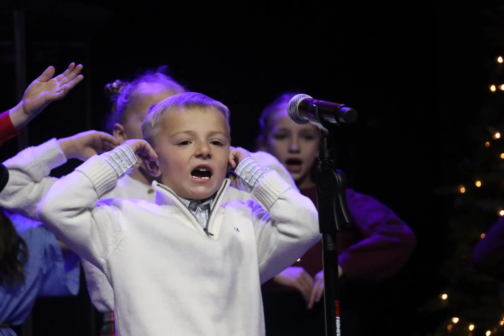 Children singing or performing on stage, some covering their ears, with a microphone in front of a dark background with lights.
