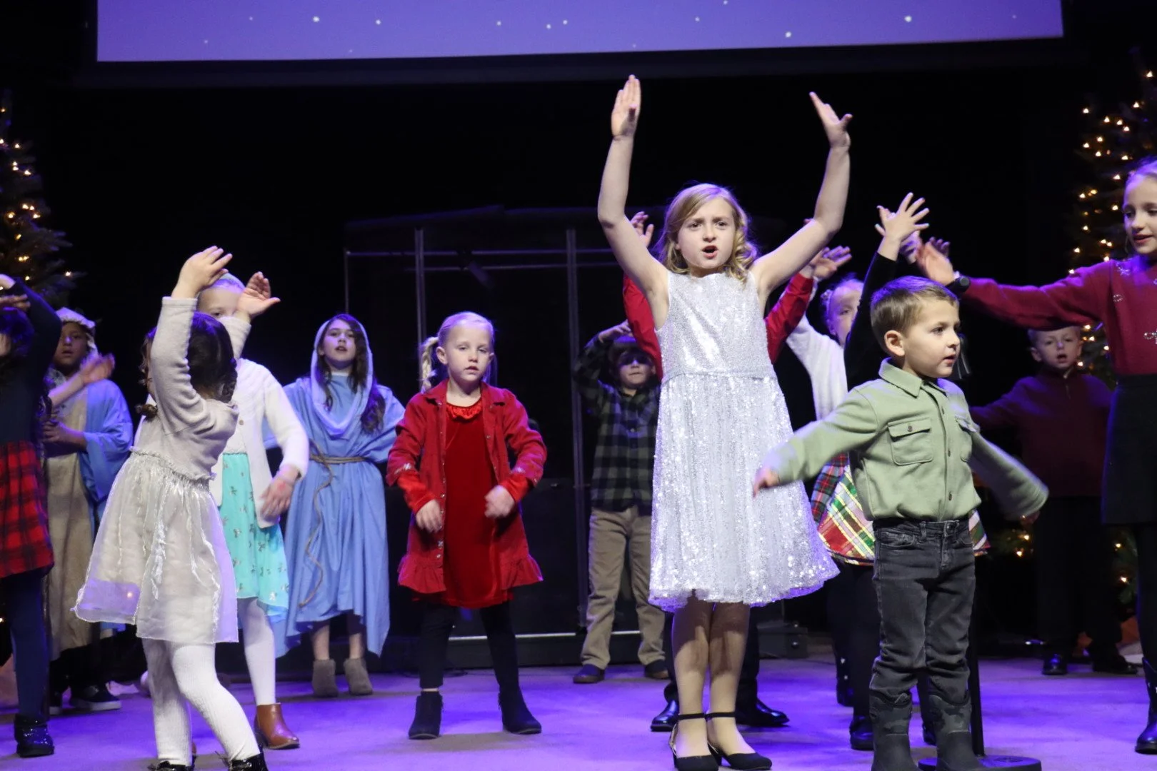 Children performing on stage in a holiday-themed performance, some with their arms raised.