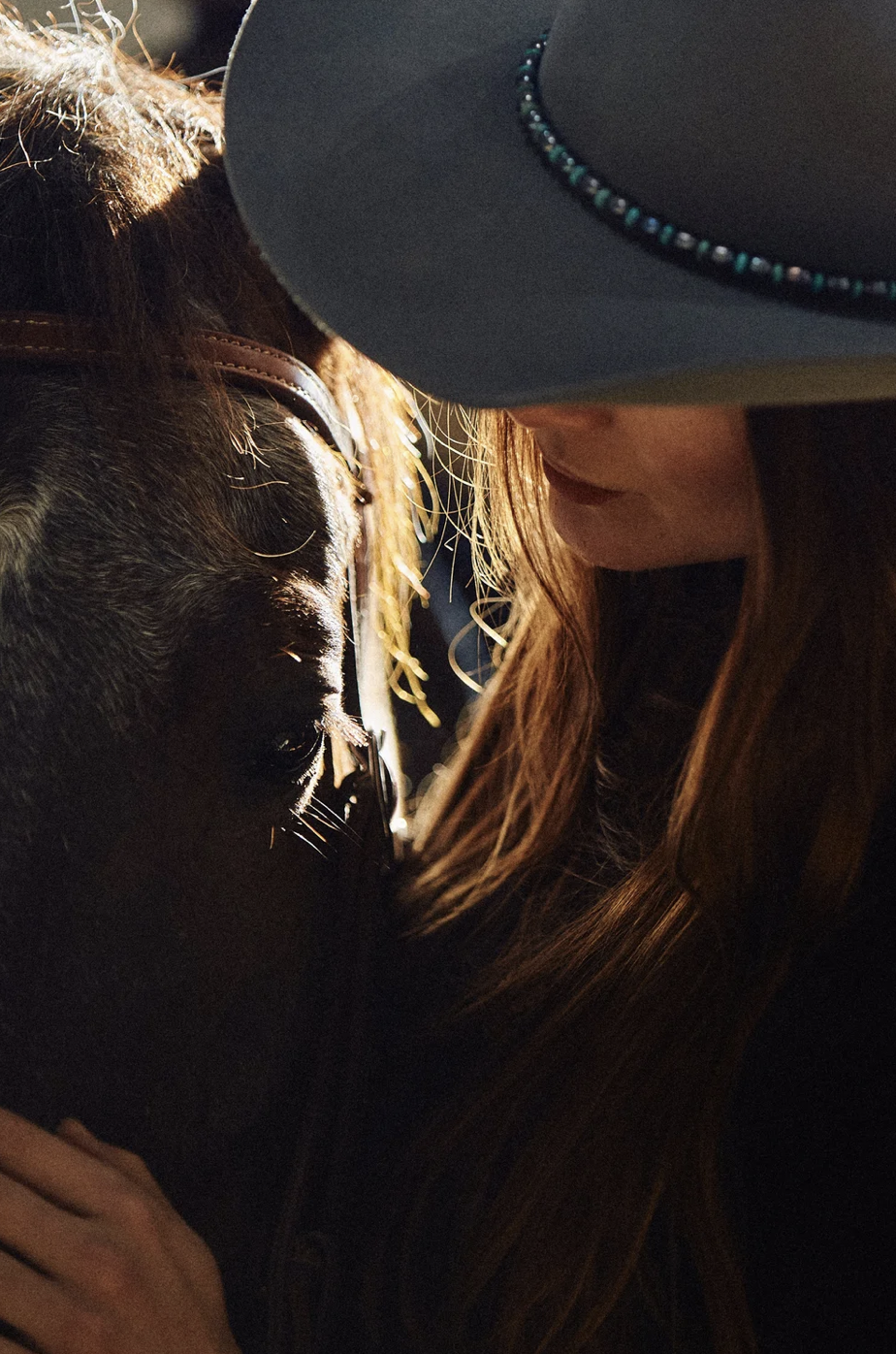 A woman with long red hair wearing a wide-brimmed hat with a beaded band is close to a black horse, gently touching its face.