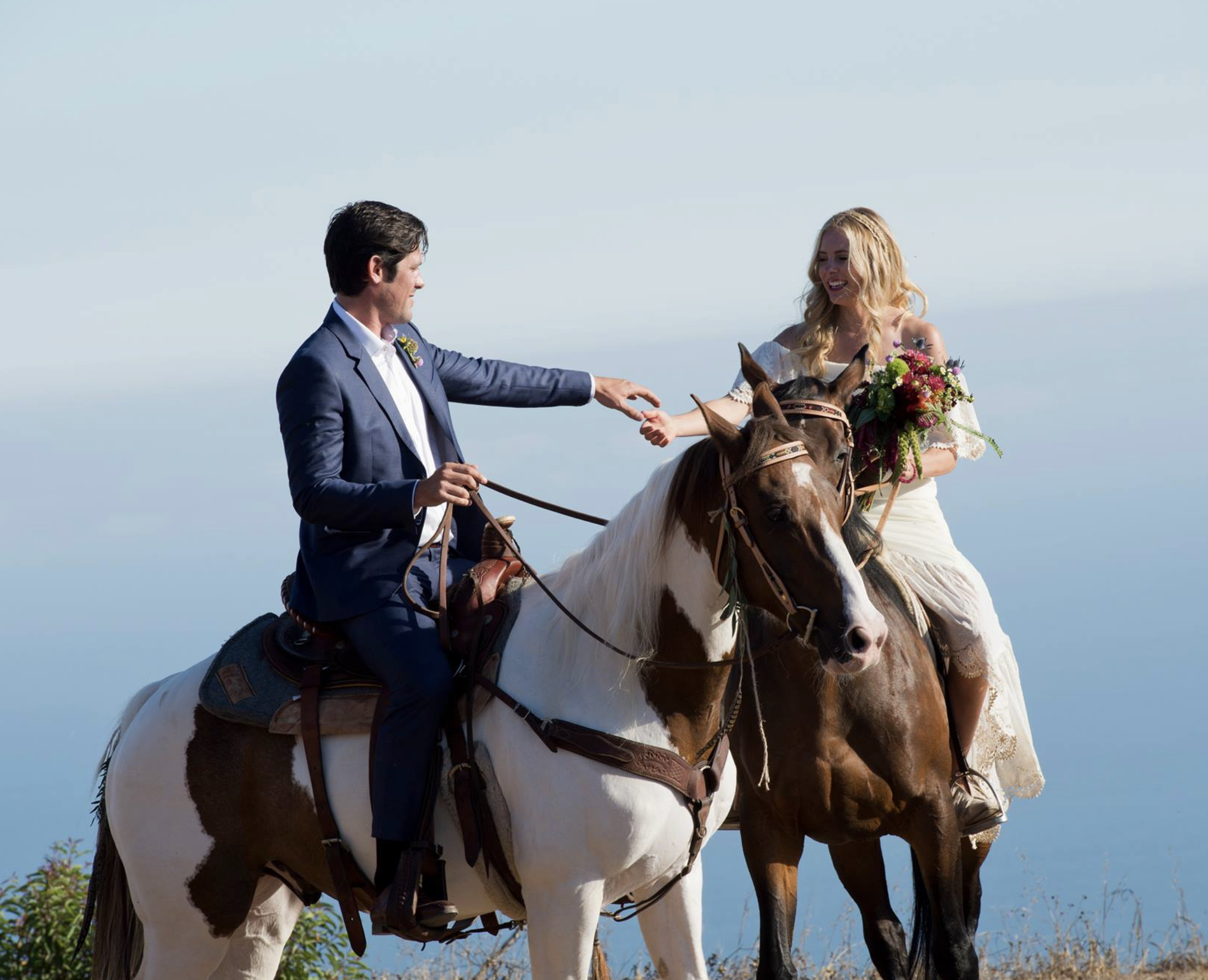A couple riding horses outdoors, with the man in a suit and the woman in a wedding dress holding a bouquet, greeting each other and smiling.