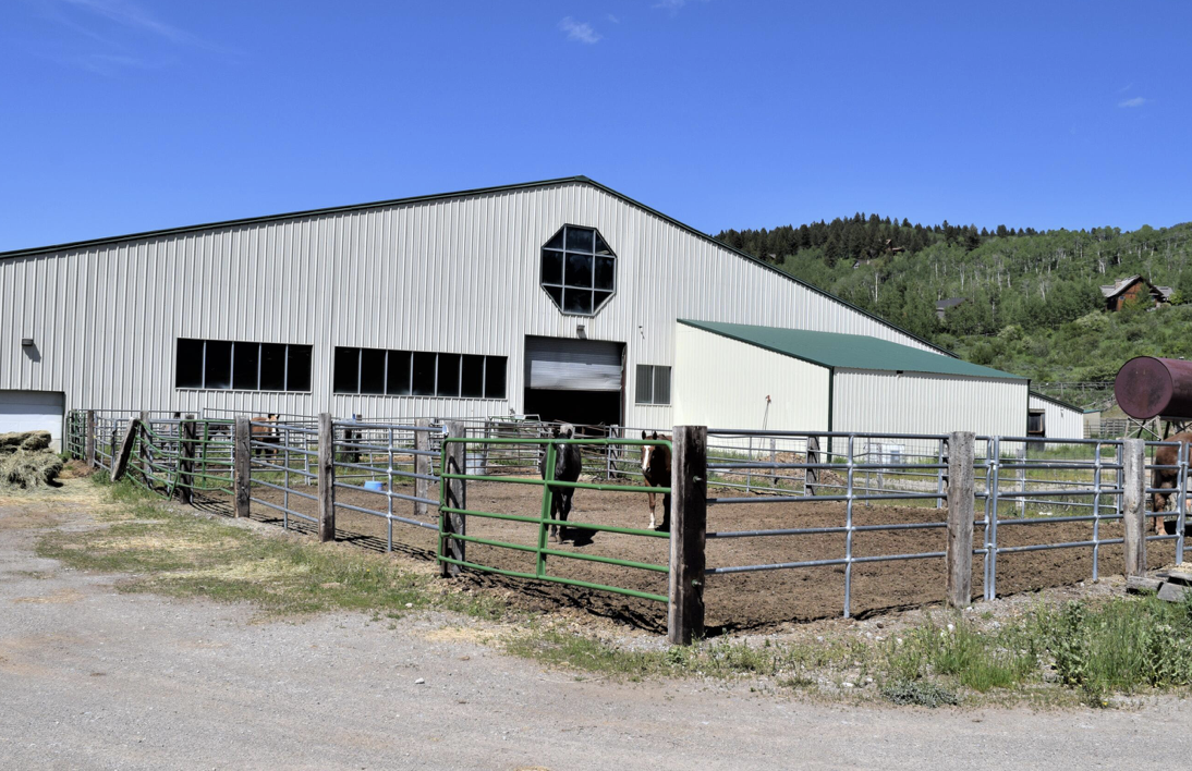 A large metal barn with an open sliding door, surrounded by a fenced area with two horses, under a blue sky in a rural setting.