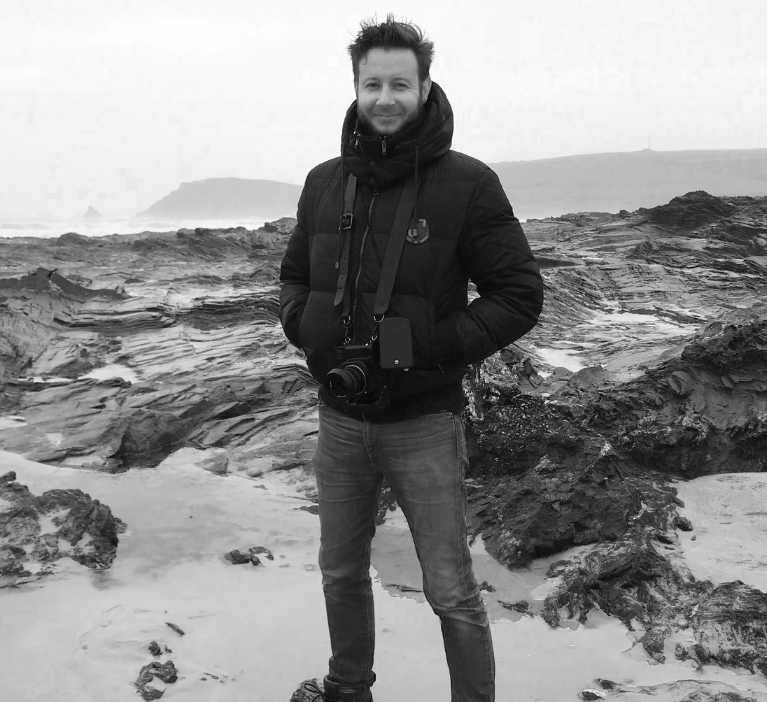 A man stands on rocky beach wearing a black jacket with camera around his neck, smiling at the camera, with ocean and misty cliffs in background.