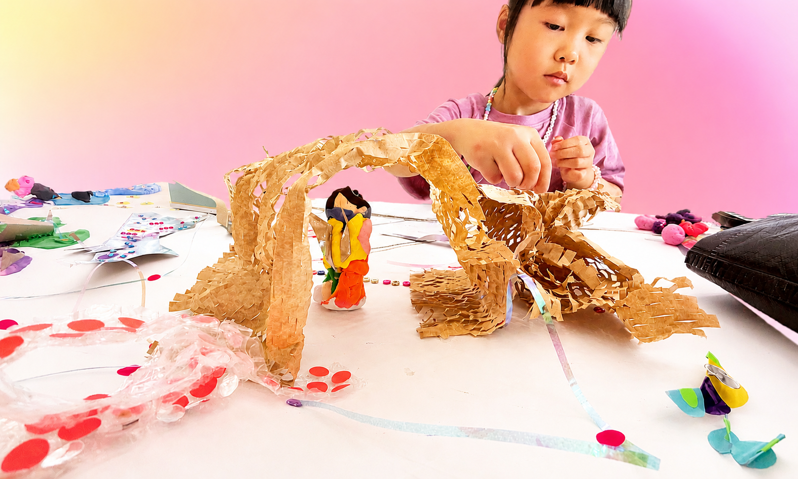 Young girl working on an art project with paper, foam, and craft supplies on a white table with pink background.