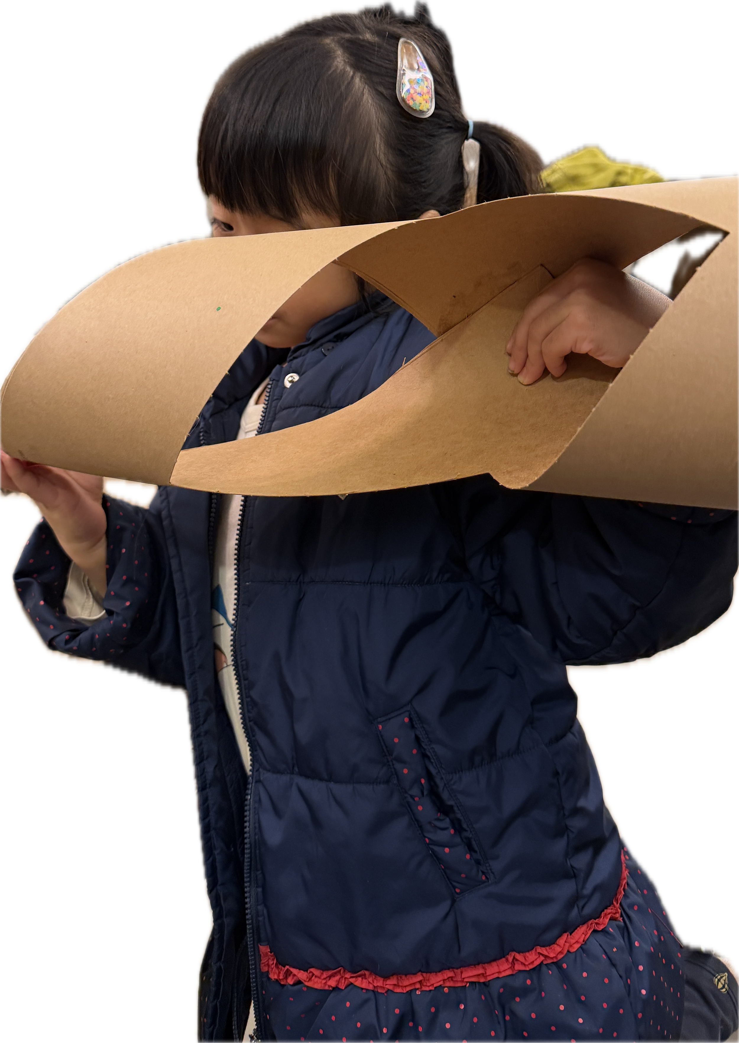 A young girl with dark brown hair in pigtails, wearing a hair clip with charms, a navy blue quilted jacket, and a dress with small red polka dots, is holding and looking at a large, curved piece of cardboard.