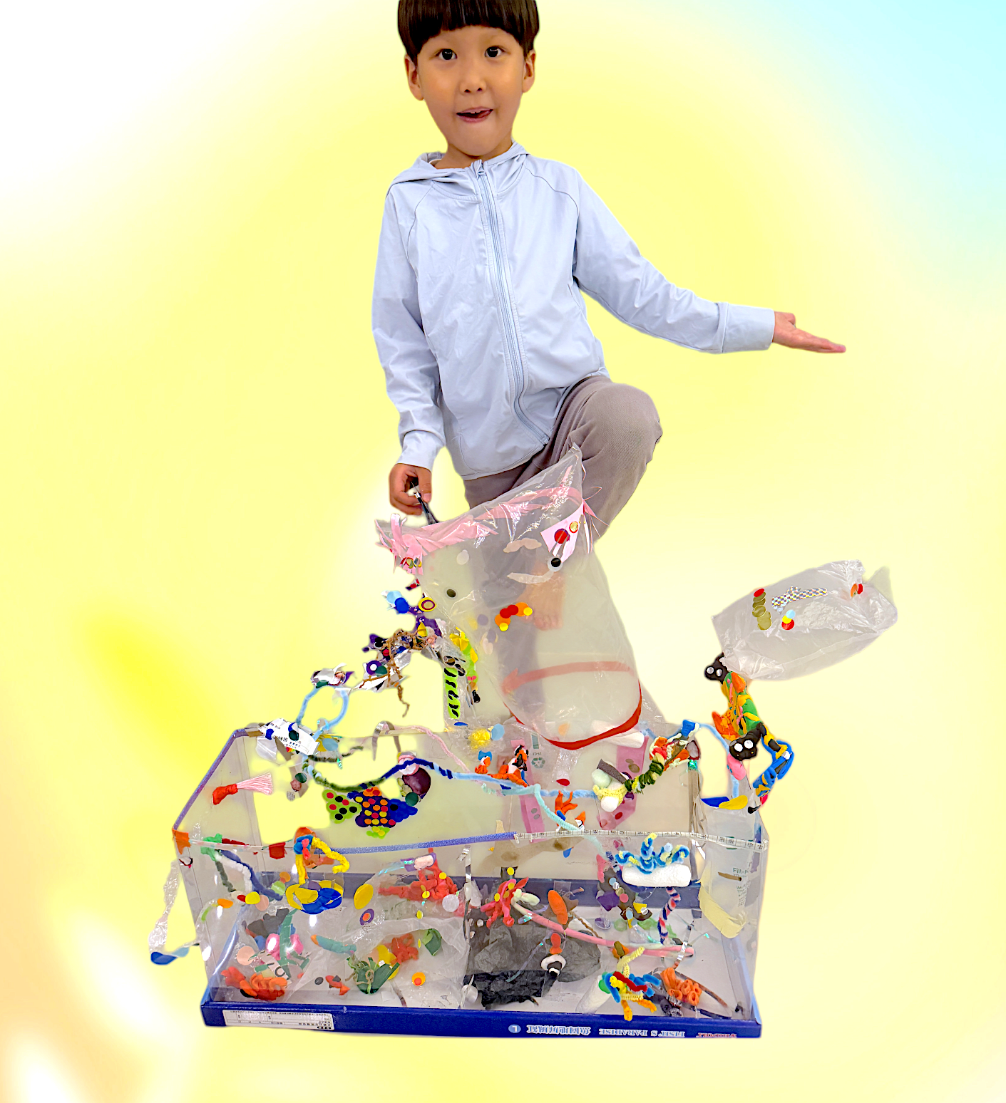 A young boy with dark hair and wearing a light gray jacket kneeling on one knee, with one arm extended to the right, standing in front of a yellow background. He is surrounded by a box filled with colorful confetti, small toys, and craft supplies.